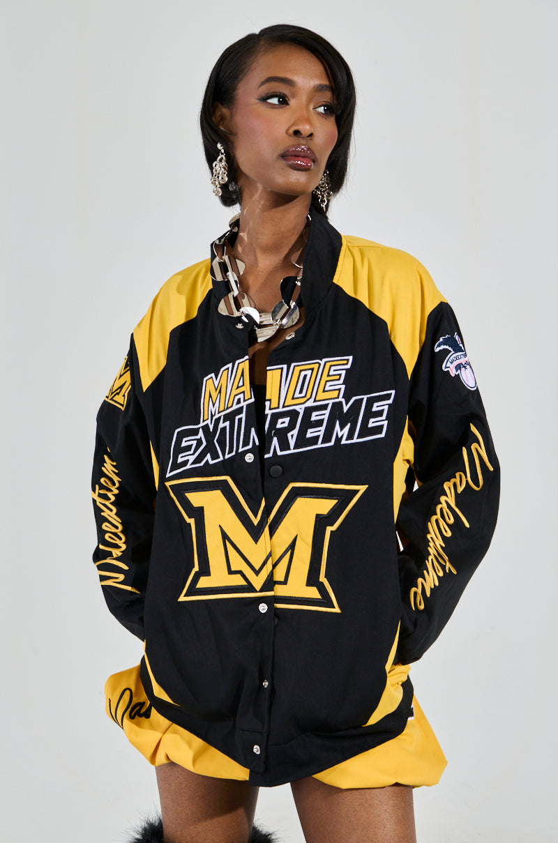A woman wears the MADE EXTREME OVERSIZED BOMBER, showcasing "Made Extreme" and an "M" logo on the front, with embroidered graphics and bold sleeve text. Styled with large hoop earrings and side-swept hair, she gazes thoughtfully to the right. 0