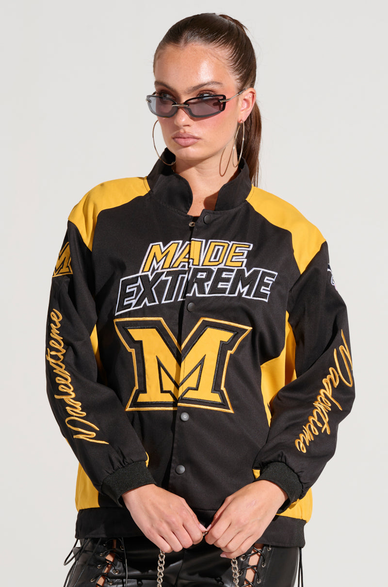 A woman wearing the MADE EXTREME OVERSIZED BOMBER sunglasses and a black and yellow varsity jacket featuring "Made Extreme" with a prominent "M" on the front. The sleeves are adorned with cursive text, which provides an embroidered graphic detail. Her long hair is pulled back as she stands against a plain background. 10