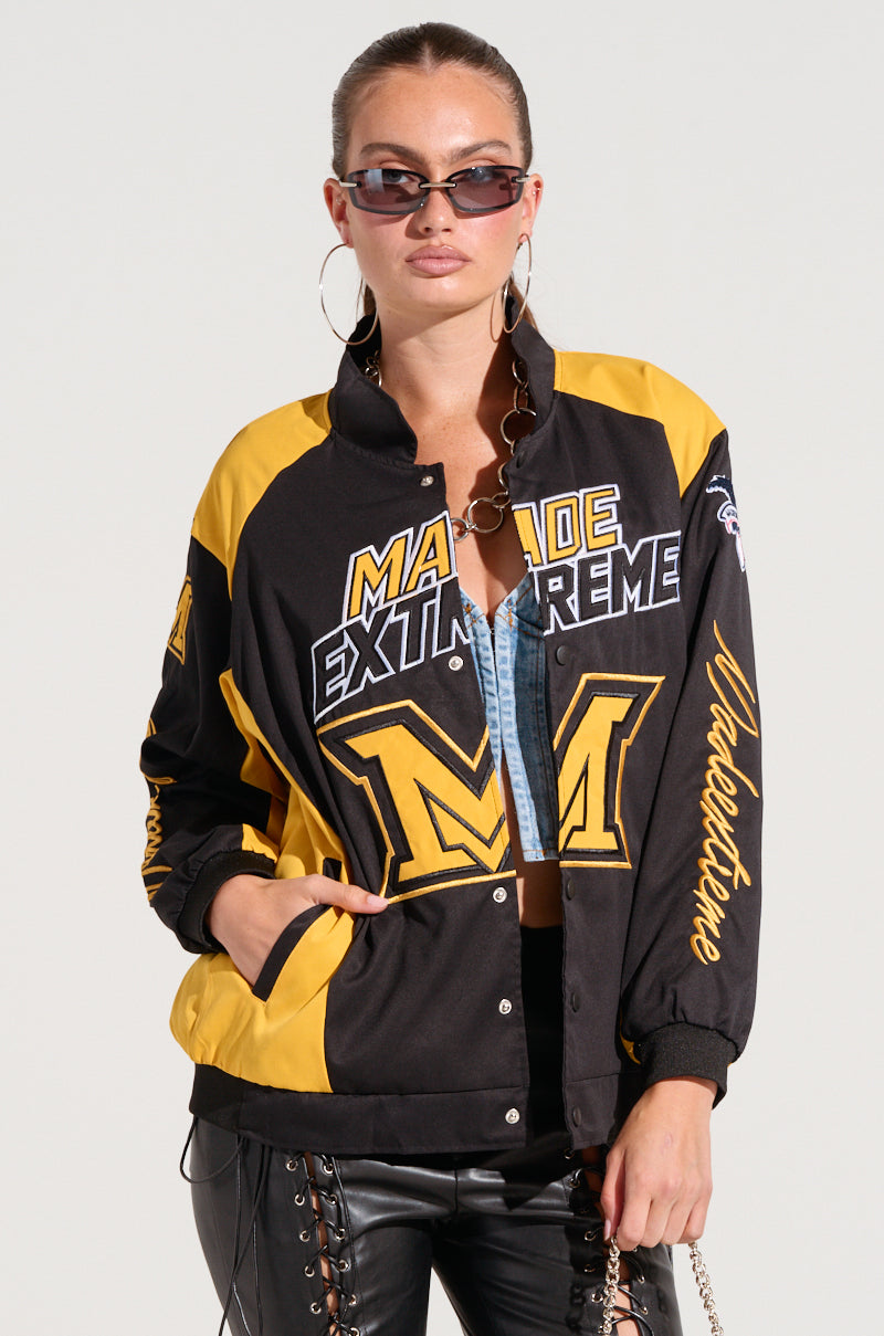 A person is sporting the MADE EXTREME OVERSIZED BOMBER, featuring black and yellow embroidered graphics with "Made Extreme" text and large "M" logos in a varsity style. They complement the look with hoop earrings, hands in pockets, standing against a plain backdrop, highlighting the trendy colorblock paneling. 11