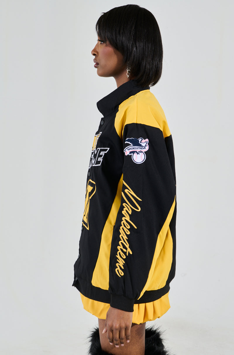 Someone with short, dark hair and earrings poses in profile against a plain backdrop, wearing the MADE EXTREME OVERSIZED BOMBER—a black and yellow varsity-style jacket featuring colorblock paneling and the phrase "Made Extreme. 2