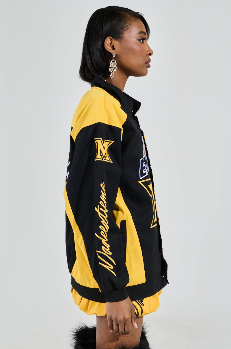 A woman is seen in profile sporting the MADE EXTREME OVERSIZED BOMBER, a black and yellow jacket with colorblock paneling and sports-inspired embroidered graphics. Her short hairstyle and large earrings complement the jacket's bold "M" and "Makestheme" design, enhancing its flair. 3