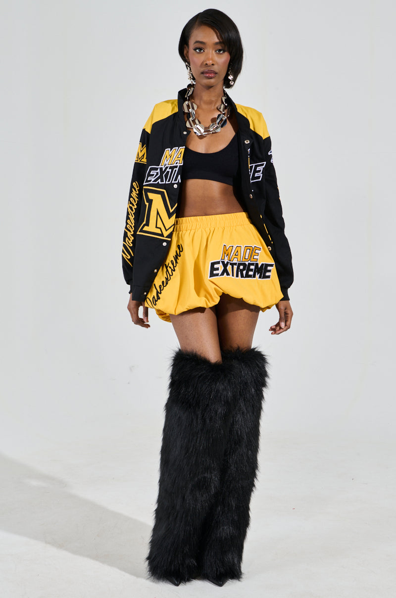 A model strides confidently on a light background, clad in the MADE EXTREME OVERSIZED BOMBER with colorblock paneling. She wears a black crop top, yellow shorts, and black furry thigh-high boots, perfectly accessorized with a chunky necklace. 6