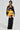 A model strides confidently on a light background, clad in the MADE EXTREME OVERSIZED BOMBER with colorblock paneling. She wears a black crop top, yellow shorts, and black furry thigh-high boots, perfectly accessorized with a chunky necklace.