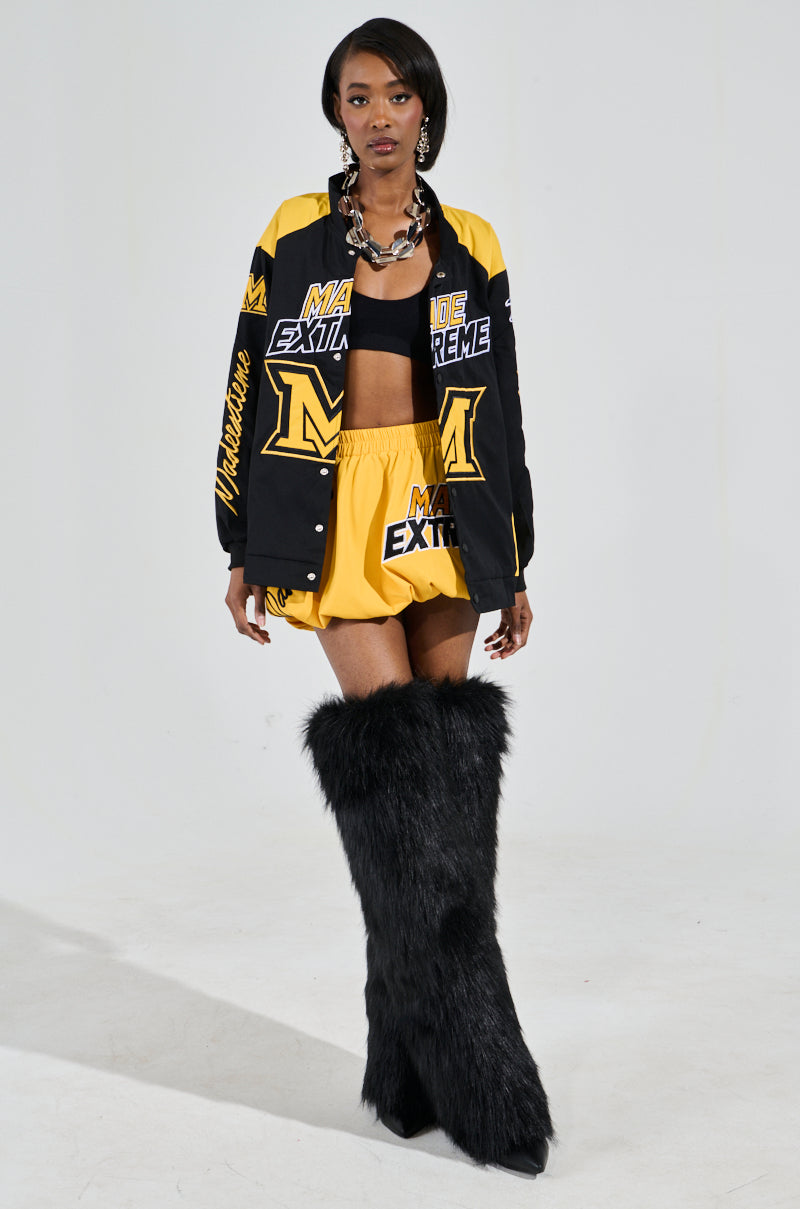 A woman models a sporty black and yellow ensemble featuring the MADE EXTREME OVERSIZED BOMBER, shorts with sports-inspired graphics, and black fuzzy knee-high boots. She completes the look with bold lettering on the outfit and a large chain necklace. 7