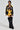 A woman models a sporty black and yellow ensemble featuring the MADE EXTREME OVERSIZED BOMBER, shorts with sports-inspired graphics, and black fuzzy knee-high boots. She completes the look with bold lettering on the outfit and a large chain necklace.