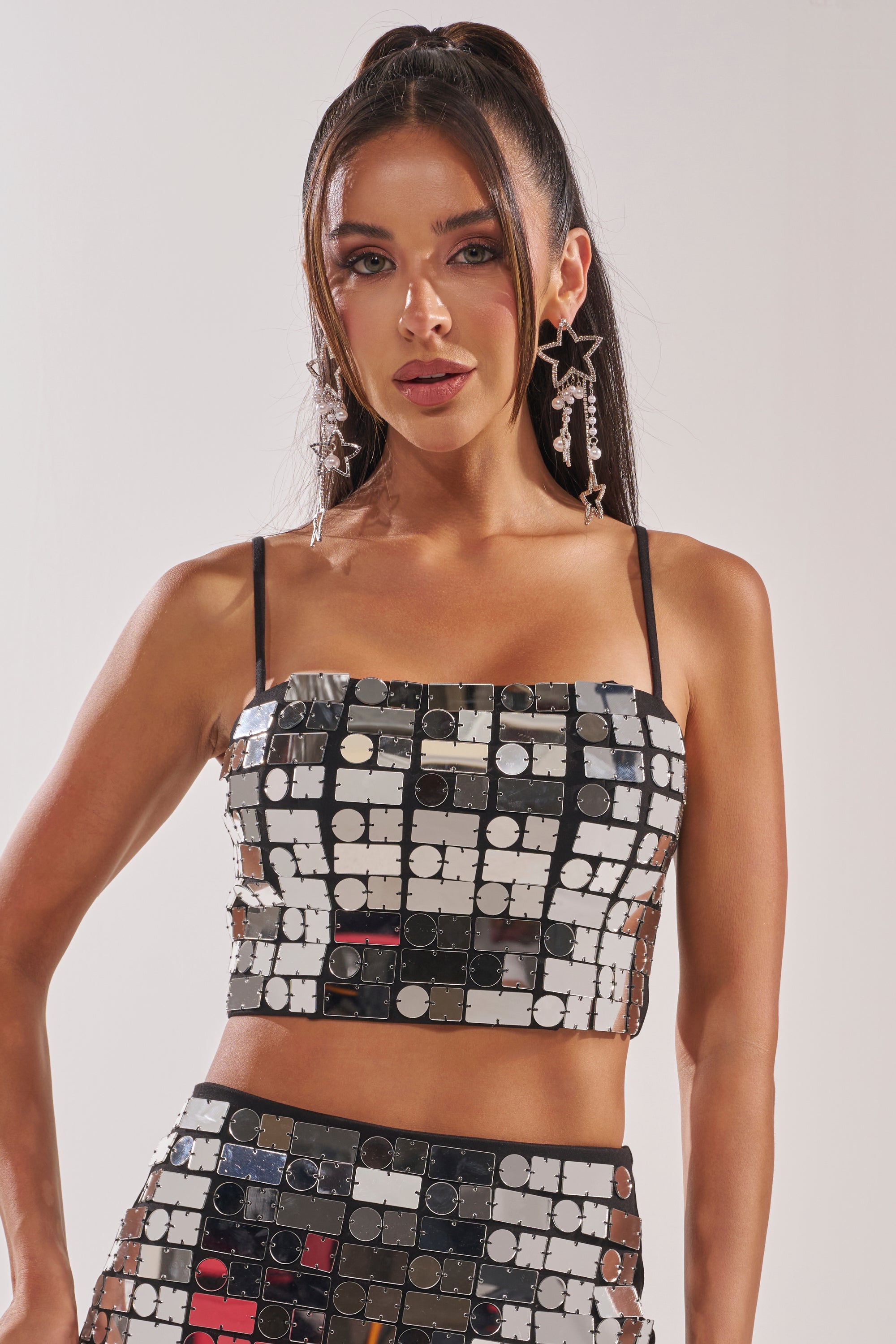 A woman with long dark hair in a high ponytail wears the REFLECTIONS BRALETTE and matching skirt, both made of geometric mirrored tiles. She accessorizes with star-shaped earrings and poses against a plain white background. 0