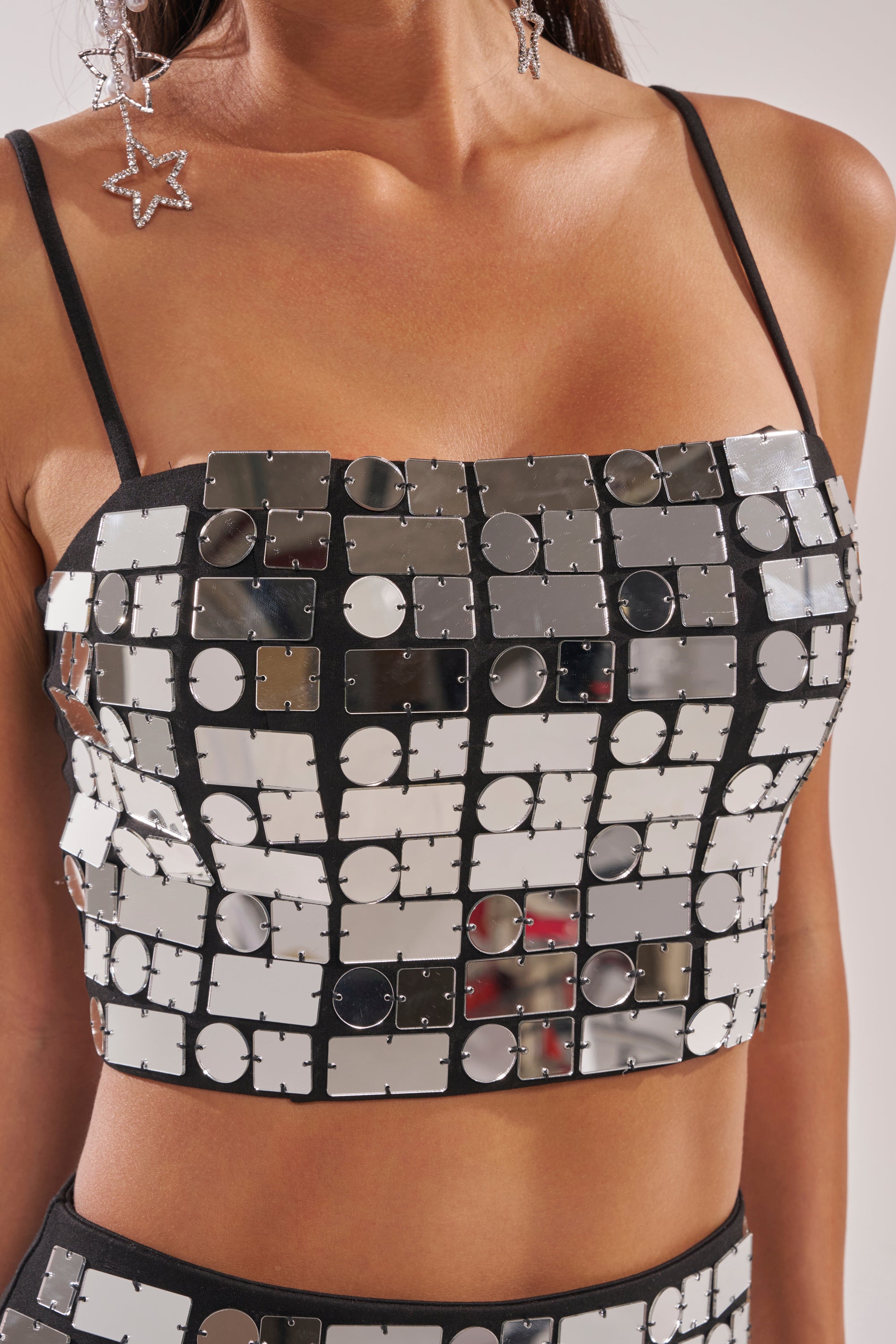 A woman models the REFLECTIONS BRALETTE, featuring shiny square and round mirrored tiles in a grid pattern. She pairs it with dangling star-shaped earrings. The photo shows her from shoulders to waist. 7