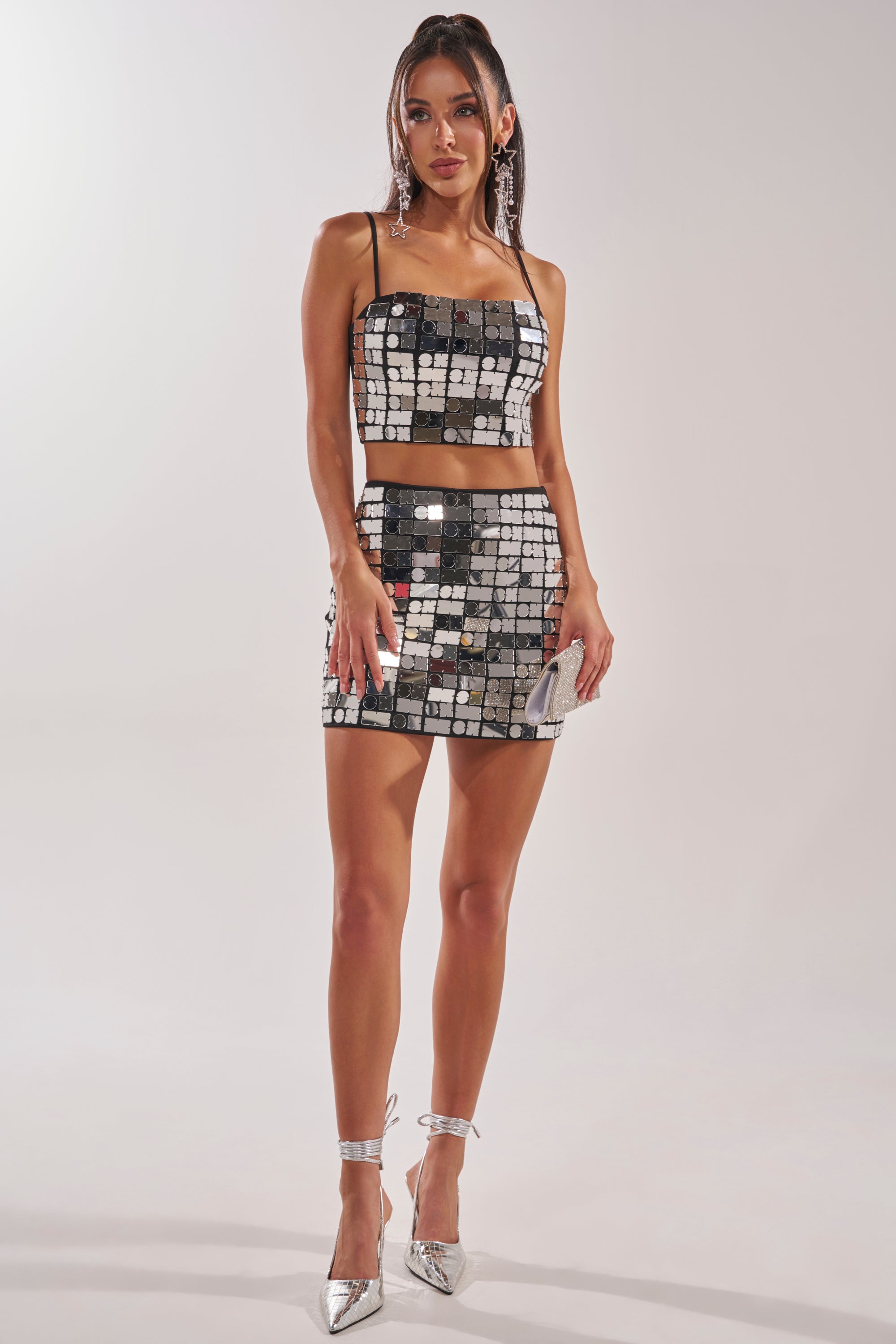 A woman wearing the REFLECTIONS MINI SKIRT and a matching mirrored crop top, styled with silver heels and a small clutch, poses against a plain light background. 0
