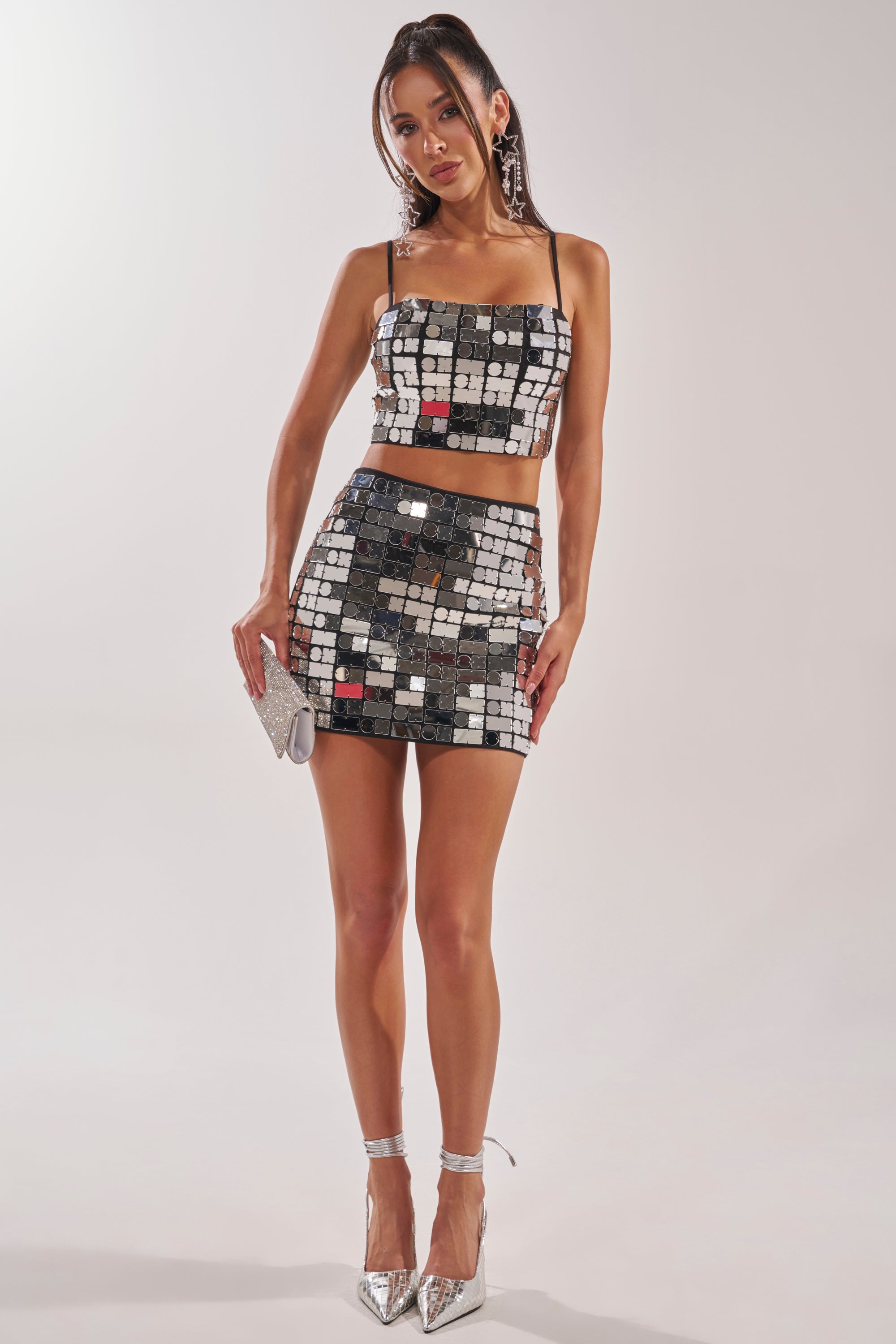 A woman poses in the REFLECTIONS MINI SKIRT, a shiny, mirrored bodycon mini paired with a matching crop top. She accessorizes with a silver clutch, silver heels, and large hoop earrings against a plain background. 7