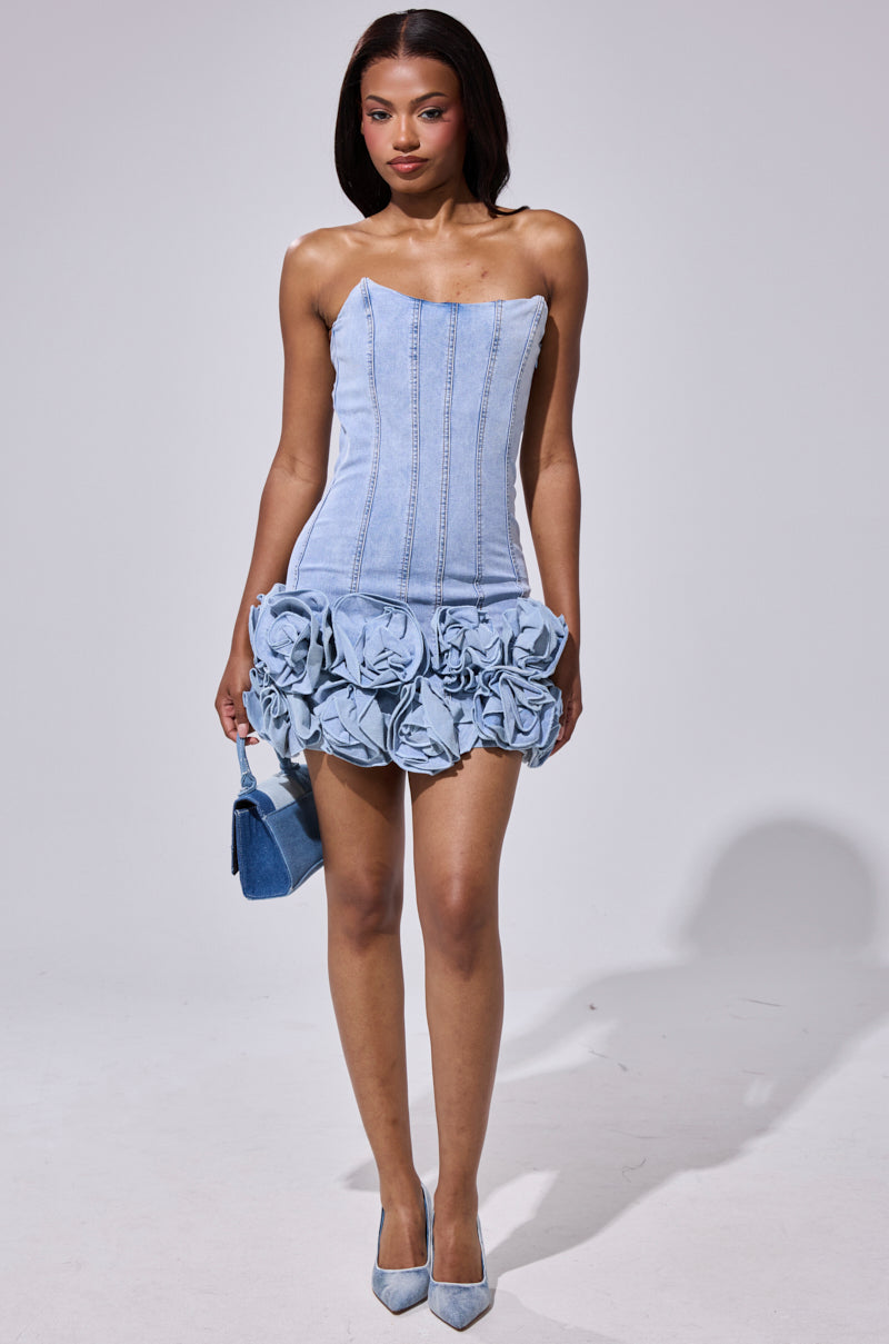 A woman wears the YOURS TRULY DENIM MINI DRESS, featuring a strapless fitted bodice, lace-up back, and large floral appliqués on the skirt. She pairs it with a small denim handbag and matching blue heels. 3