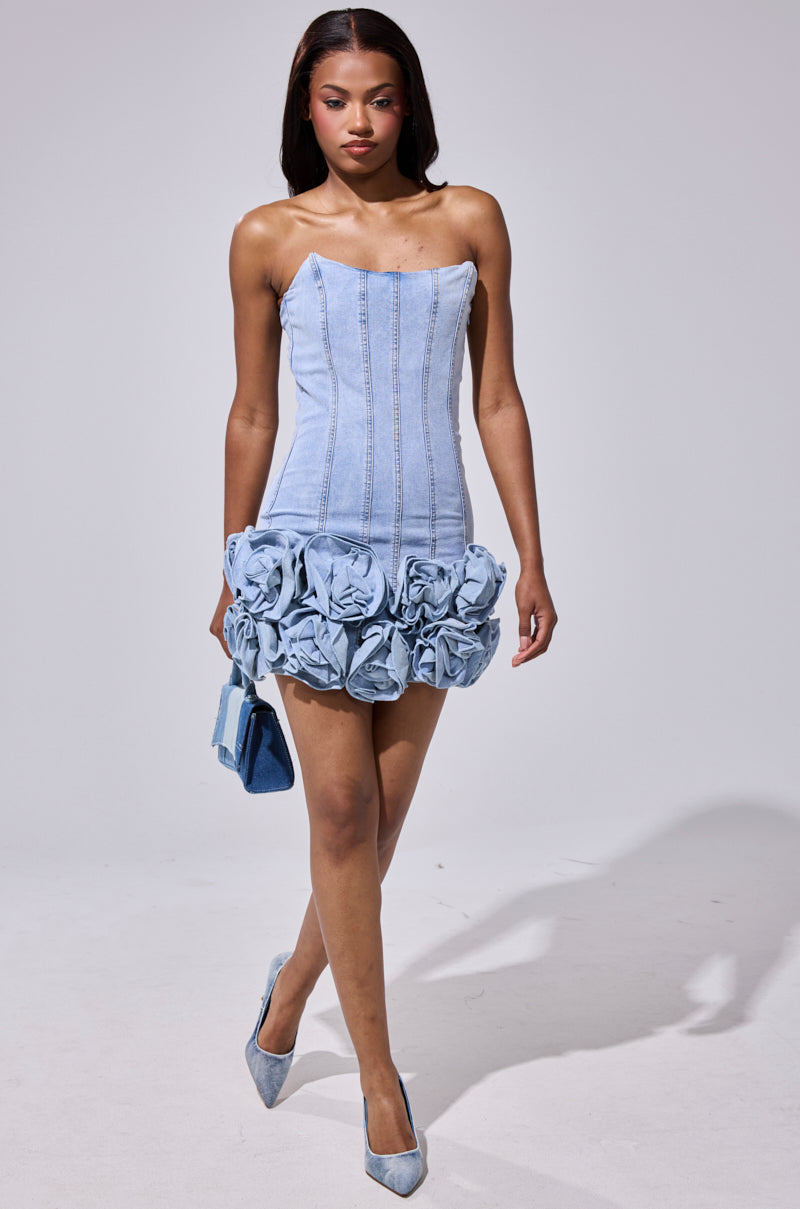 Wearing the YOURS TRULY DENIM MINI DRESS—strapless, light blue, with a lace-up back and denim rosettes—a woman carries a matching bag and dons light blue heels as she walks confidently against a plain background. 5