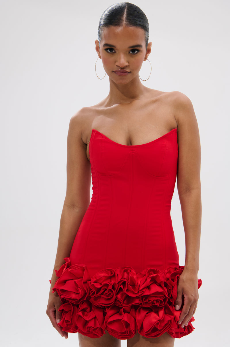 A woman stands against a plain light background wearing the ROSES ARE RED CORSET MINI DRESS, a strapless mini dress with ruffled flower detail along the hem. Her hair is pulled back and she accessorizes with large hoop earrings.