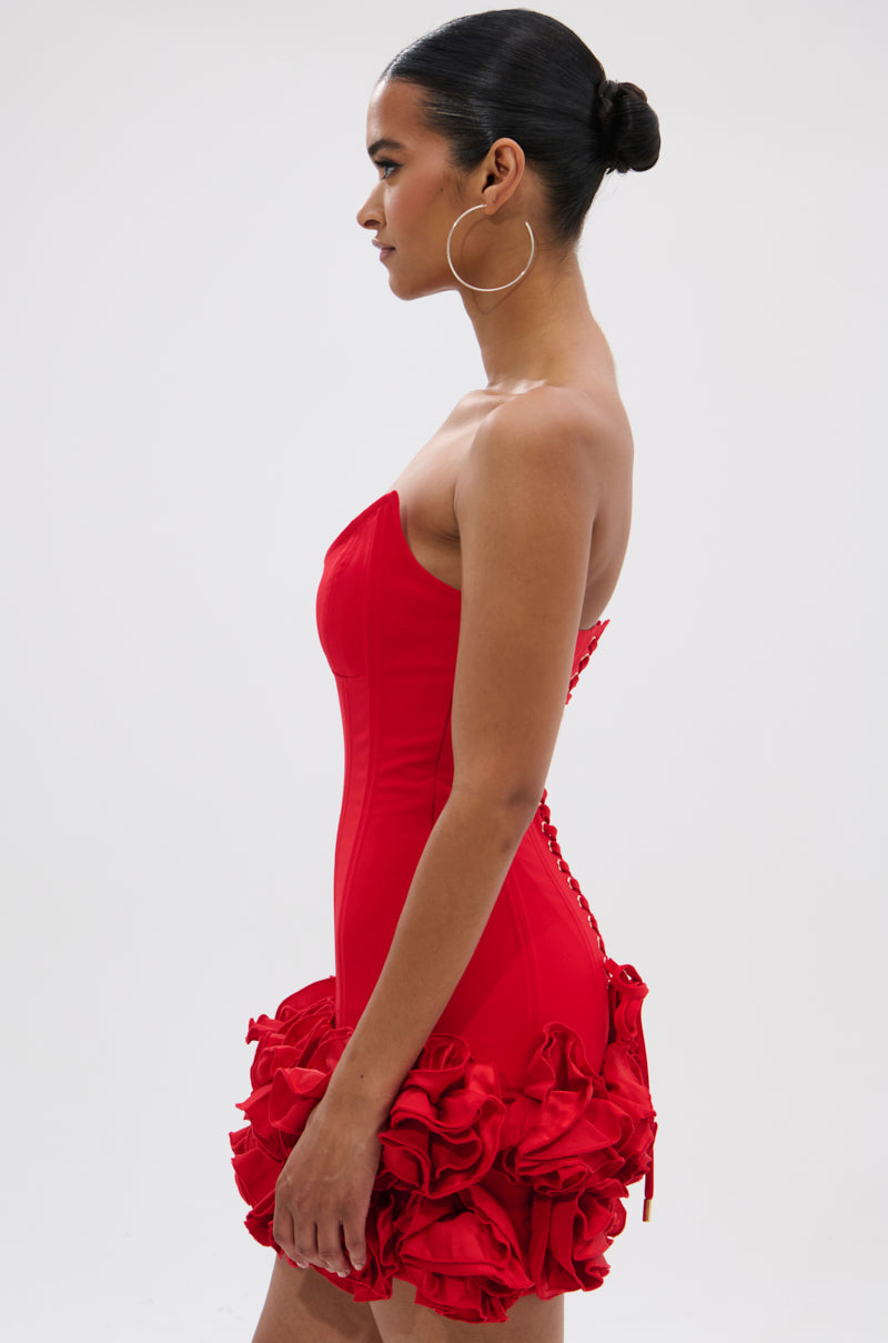 Woman in profile wearing the ROSES ARE RED CORSET MINI DRESS, a strapless mini dress with ruffled skirt detailing. She has her hair styled in a sleek bun and accessorizes with large hoop earrings, standing against a plain white background.