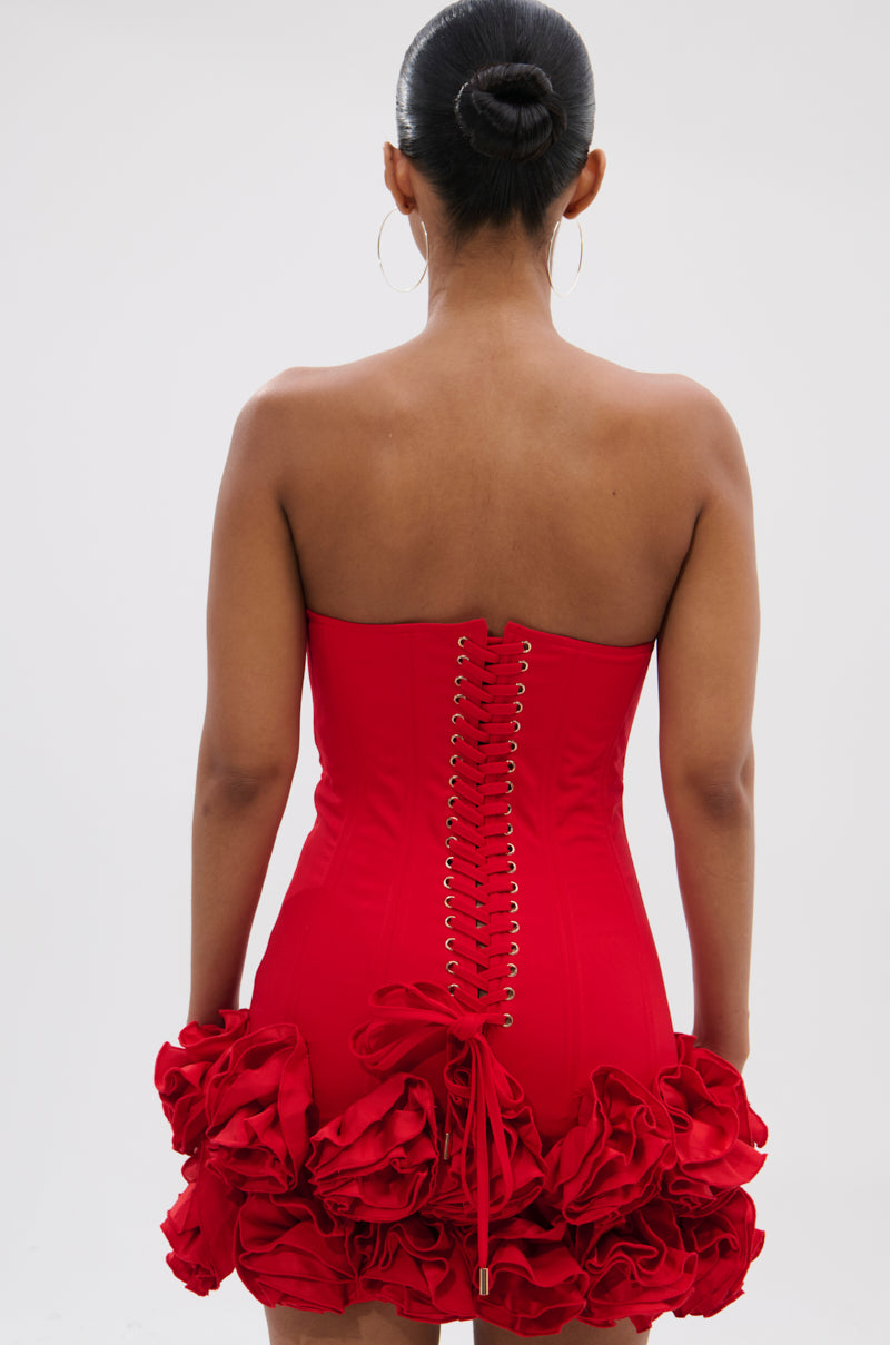 A woman with her hair in a bun models the ROSES ARE RED CORSET MINI DRESS, a vibrant red strapless mini featuring a lace-up back and ruffled hem, against a plain white background.