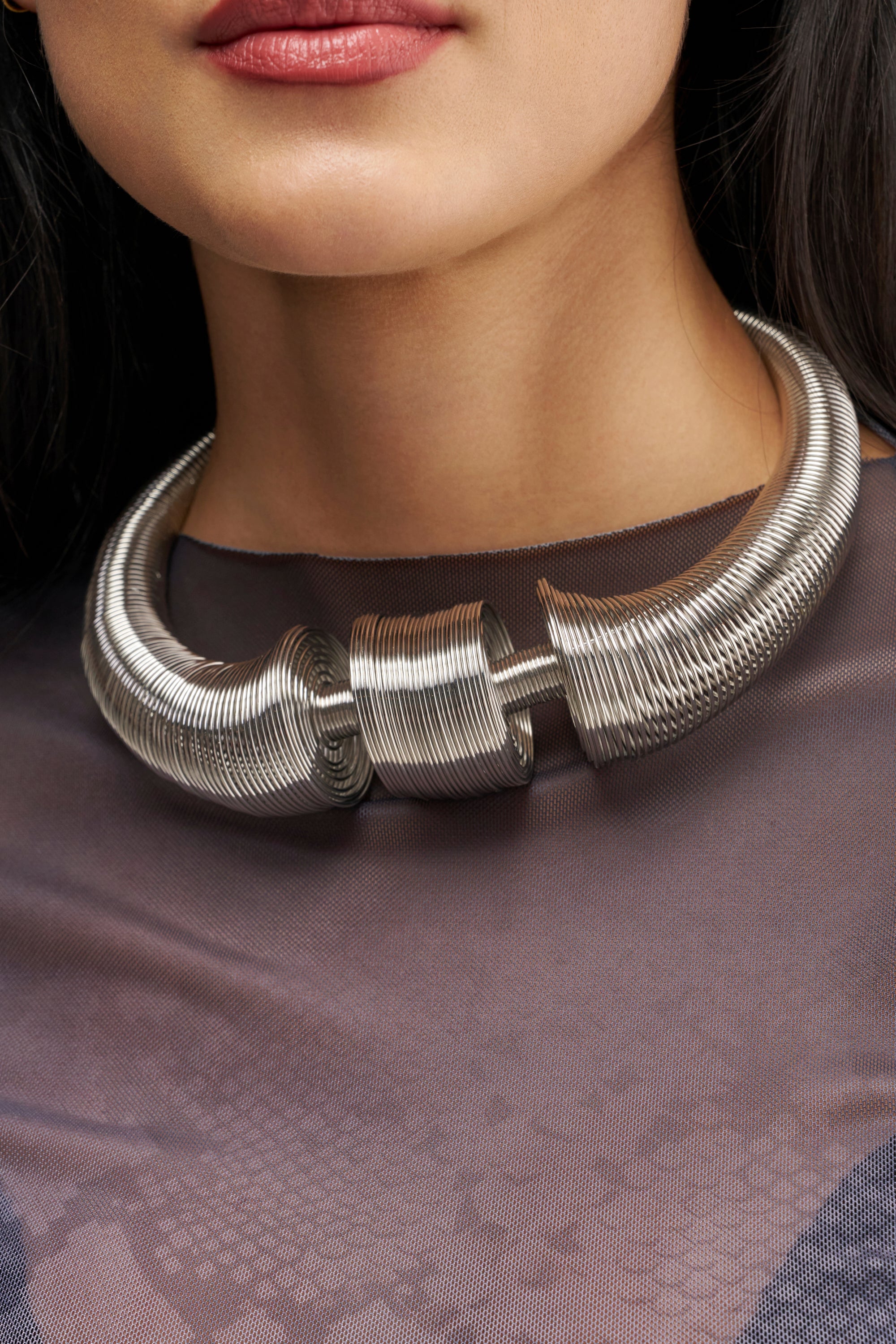 A woman wearing the SCULPTED COIL COLLAR IN SILVER over a sheer, dark top. Only the lower half of her face and neck are visible.
