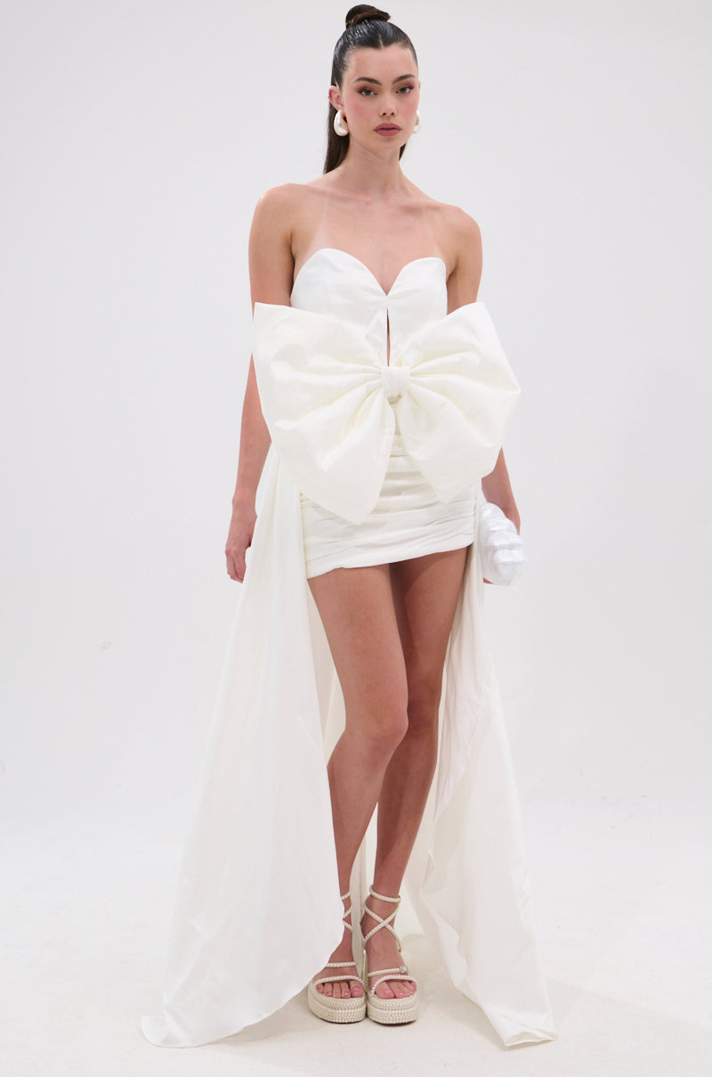 A woman wears the SHE IS THE MOMENT MINI DRESS WITH TRAIN IN WHITE, a strapless mini with a sweetheart neckline, front bow detail, and long high-low train. She pairs it with white earrings and platform sandals against a plain white background. 1