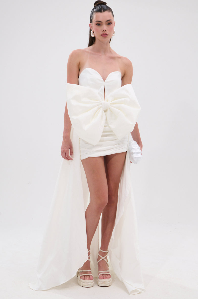 A woman wears the SHE IS THE MOMENT MINI DRESS WITH TRAIN IN WHITE, a strapless mini dress featuring a large front bow and dramatic high-low back panels. She pairs it with platform sandals and poses against a plain white background, hair in a high ponytail. 2