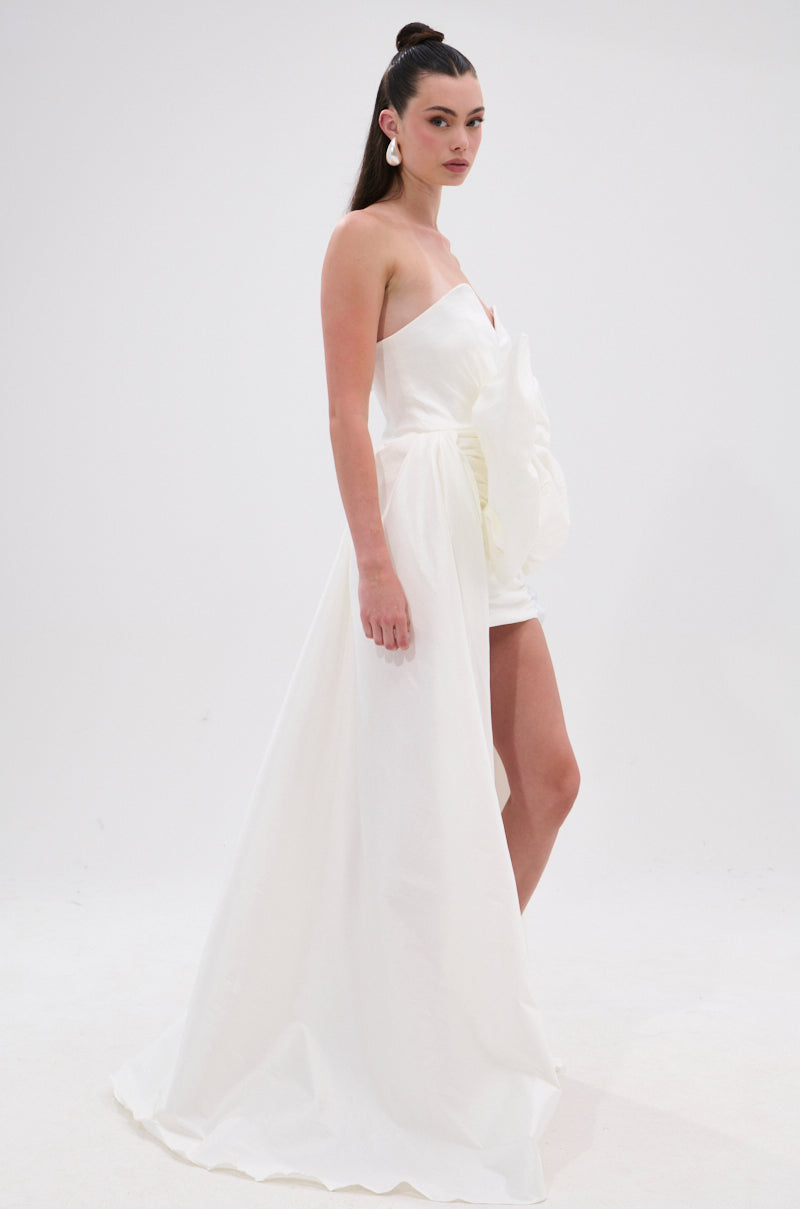 A woman with a high ponytail wears the SHE IS THE MOMENT MINI DRESS WITH TRAIN IN WHITE, a strapless mini dress with a fitted skirt and dramatic high-low train. She stands sideways against a plain white background, wearing statement earrings. 3