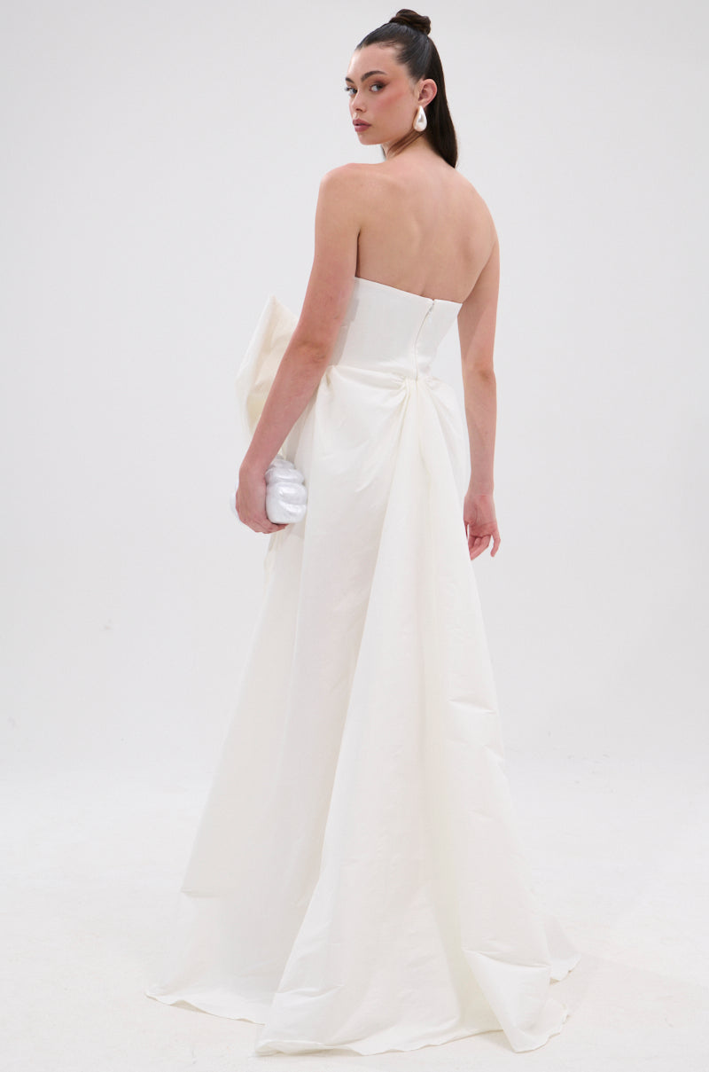 Wearing the SHE IS THE MOMENT MINI DRESS WITH TRAIN IN WHITE, a woman stands slightly turned away, holding a white clutch. The strapless mini dress features a sweetheart neckline and dramatic back bow. She sports long, dark hair in a high ponytail and statement earrings. 4