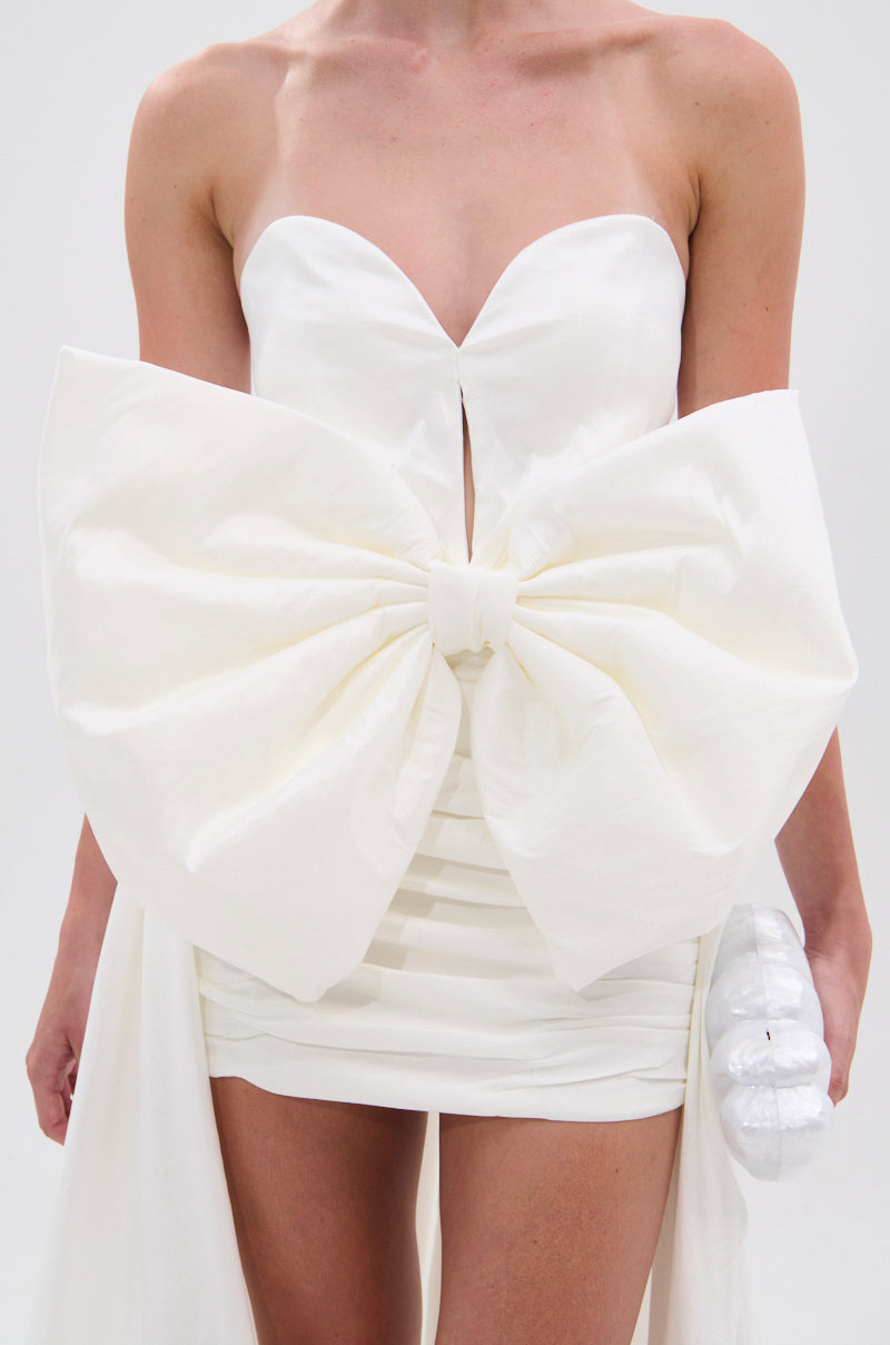 A person wears the SHE IS THE MOMENT MINI DRESS WITH TRAIN IN WHITE, a strapless mini dress with a dramatic front bow, sweetheart neckline, fitted silhouette, elegant high-low train, and holds a small white clutch. 6