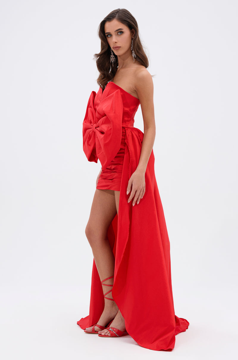 A woman poses against a white background in the SHE IS THE MOMENT MINI DRESS WITH TRAIN IN RED, featuring a strapless neckline, front bow detail, ruched mini skirt, and a dramatic long train. She completes the look with strappy heels and statement earrings.
