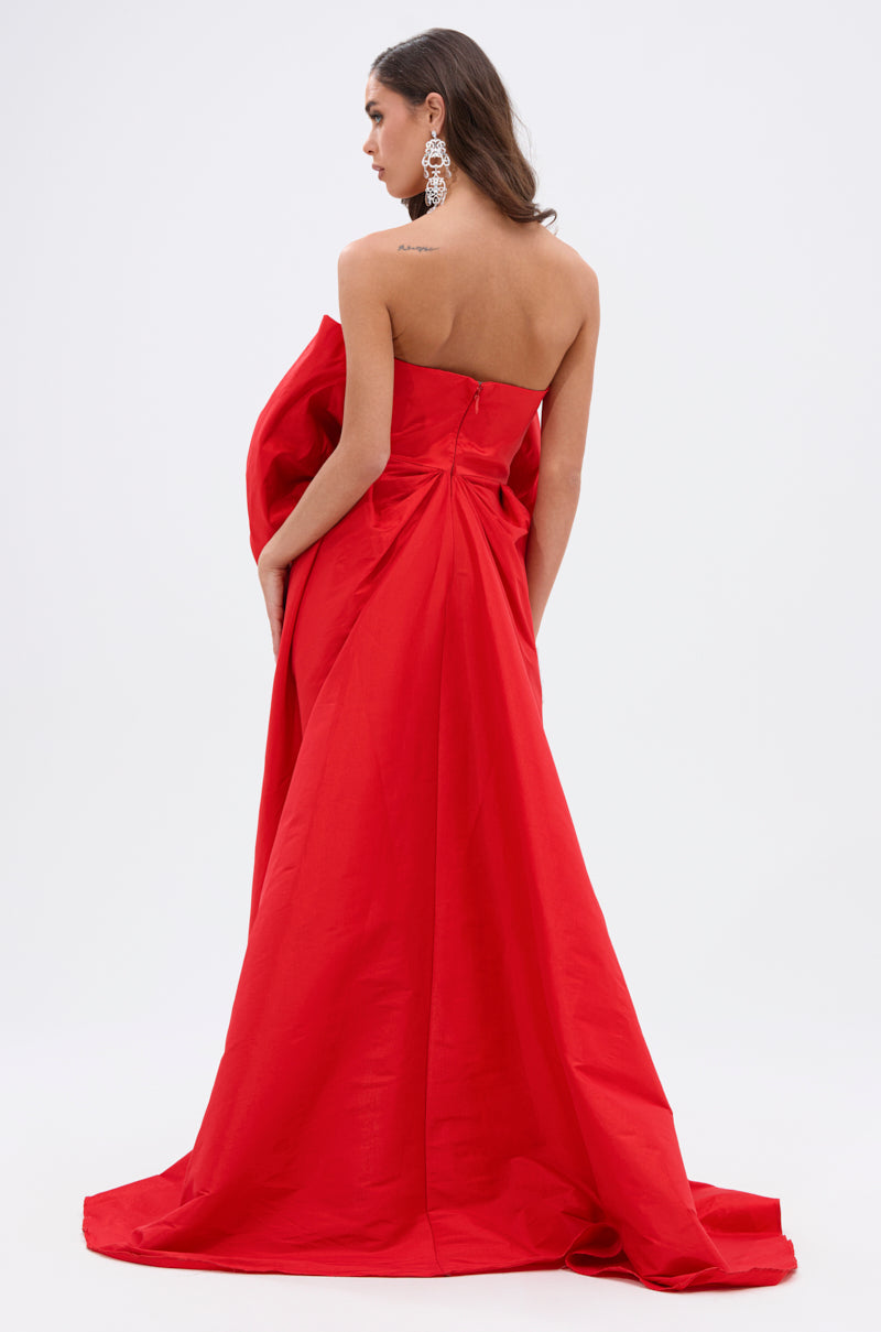 A woman with long hair, large earrings, and an upper back tattoo is seen from behind wearing the SHE IS THE MOMENT MINI DRESS WITH TRAIN IN RED, featuring a strapless neckline and dramatic draped train. Plain white background.