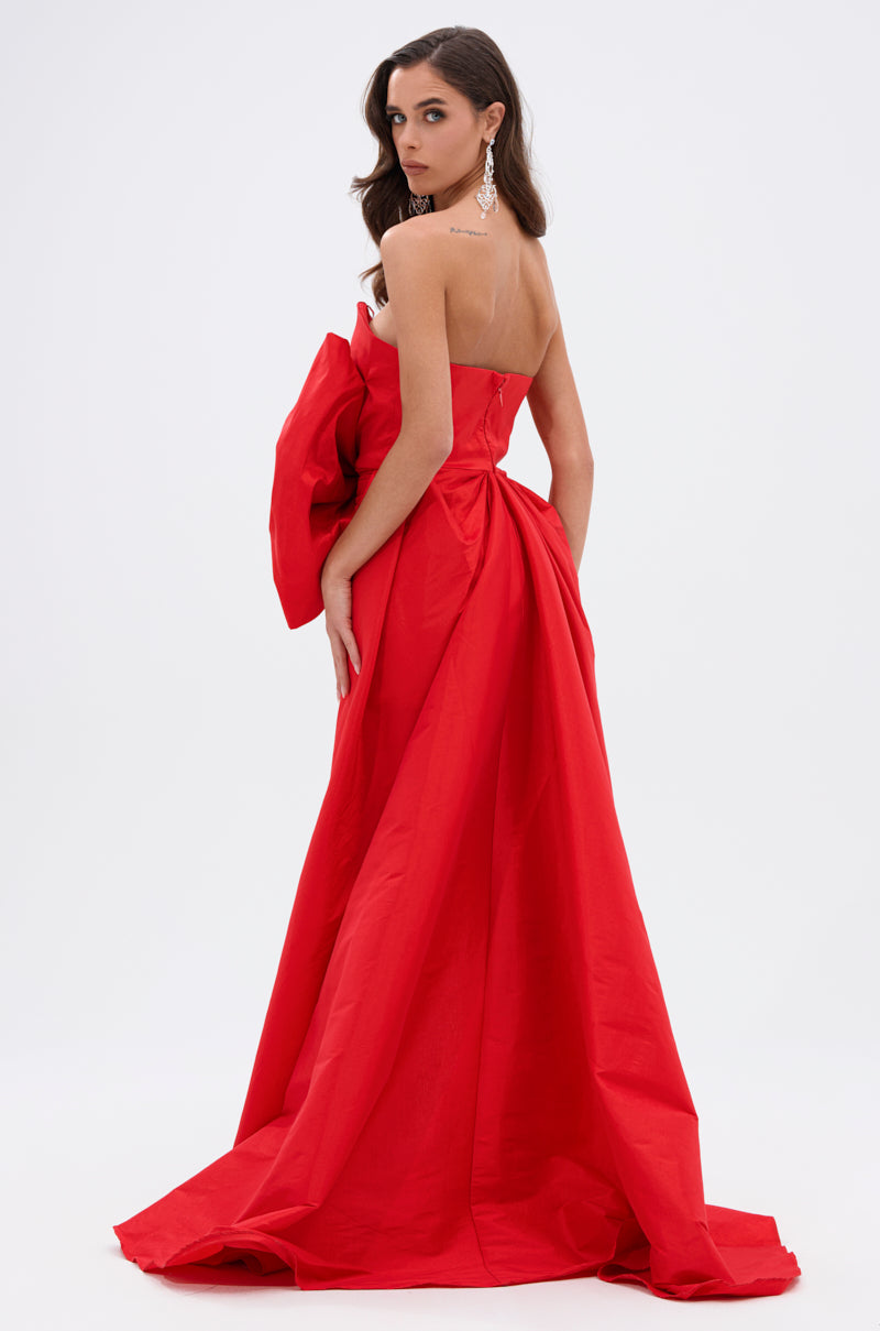 A woman poses sideways in the SHE IS THE MOMENT MINI DRESS WITH TRAIN IN RED, featuring a strapless neckline and dramatic back bow. She looks over her shoulder, with long brown hair and dangling earrings, against a plain white background.
