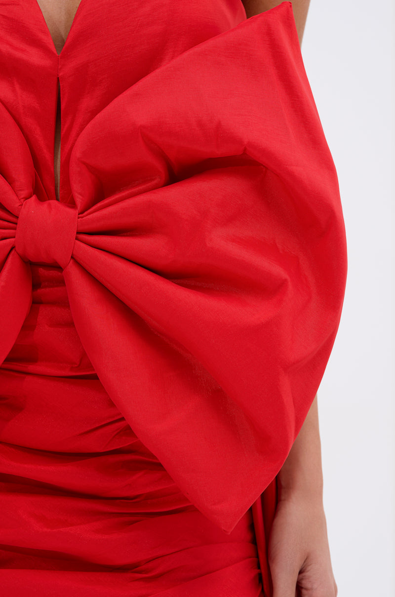 Close-up of a person wearing the SHE IS THE MOMENT MINI DRESS WITH TRAIN IN RED, featuring a strapless neckline and dramatic structured bow at the chest. The shiny fabric highlights the striking upper torso and arm design.