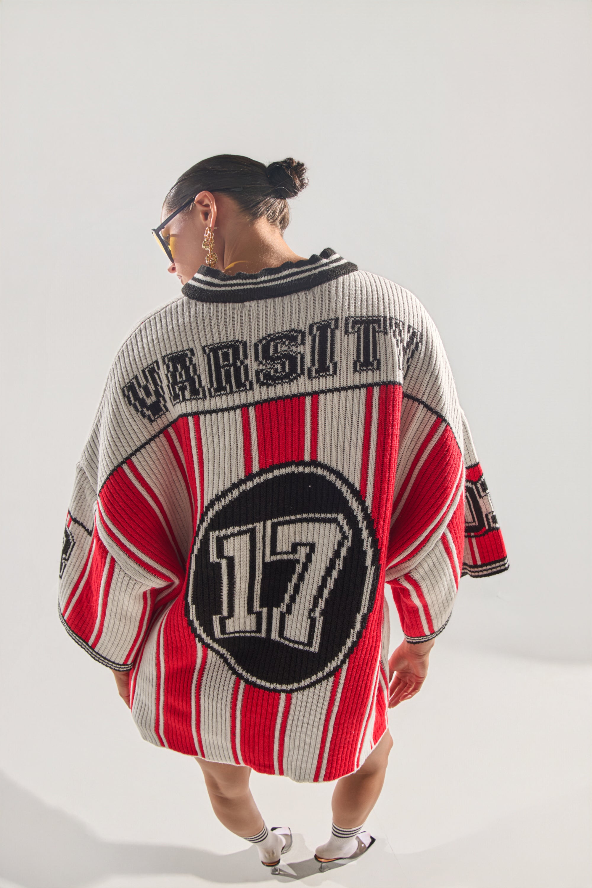 A person with dark hair in a bun wears the THROW SUM MO KNIT JERSEY TOP, an oversized varsity sweater featuring bold red, white, and black stripes and "VARSITY 17" on the back, standing against a plain light background. 2