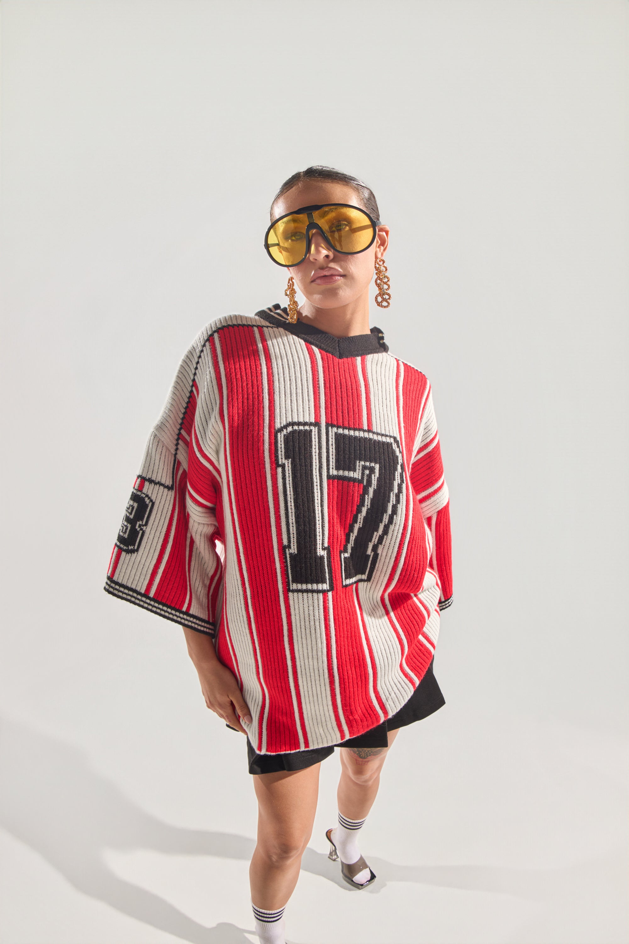 A person wears the THROW SUM MO KNIT JERSEY TOP—an oversized varsity sweater with red, white, and black stripes and the number 17—paired with large yellow sunglasses, hoop earrings, black shorts, and socks with sandals against a light background. 3