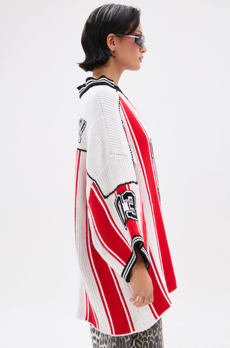 A person with short dark hair and sunglasses models the THROW SUM MO KNIT JERSEY TOP— a red, white, and black varsity-style sweater with "13" on the sleeve—paired with a leopard print skirt against a plain white background.