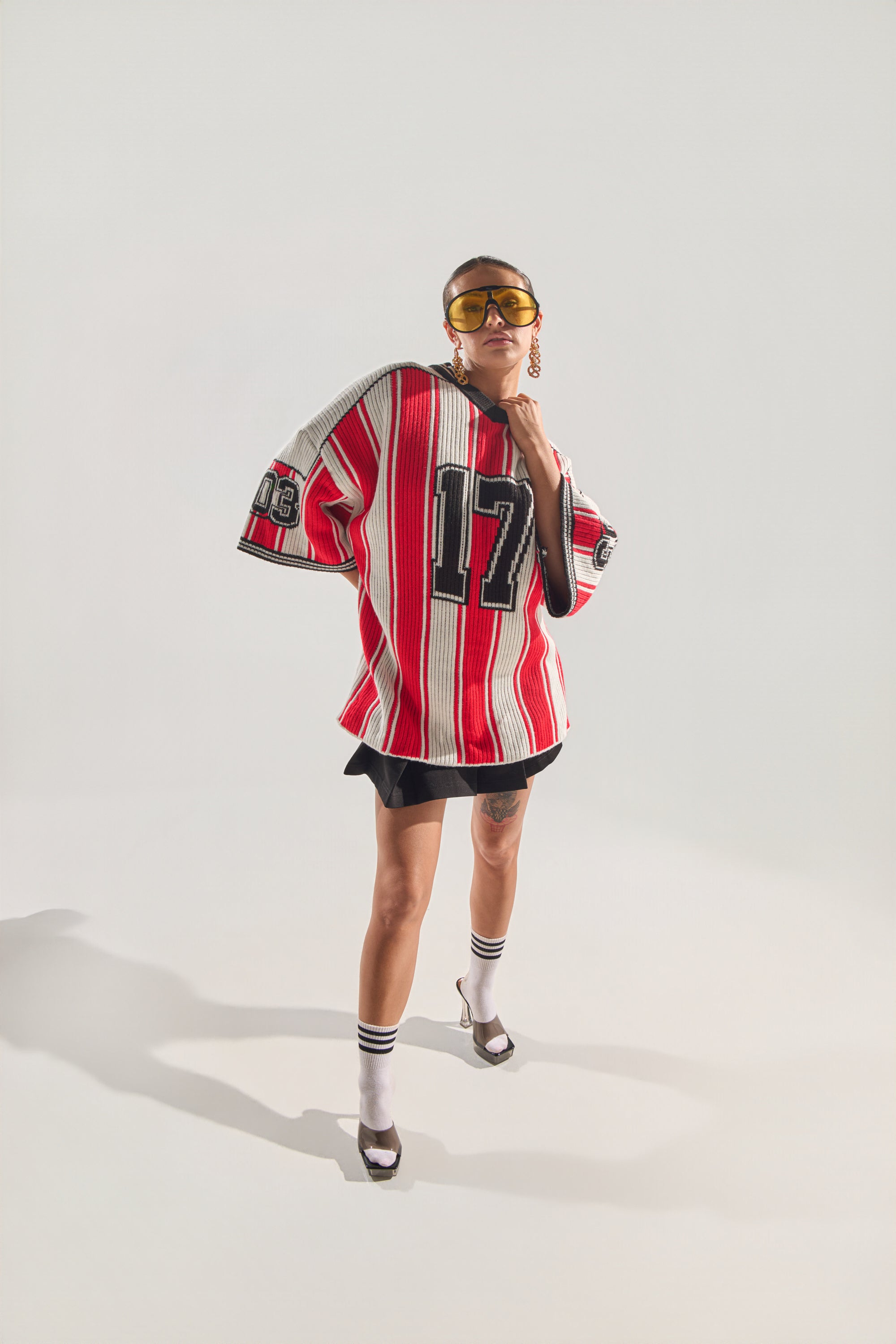 A person wearing yellow sunglasses, a black skirt, white socks, open-toe heels, and the THROW SUM MO KNIT JERSEY TOP with red and white stripes and the number 17 poses confidently against a plain light background. 4
