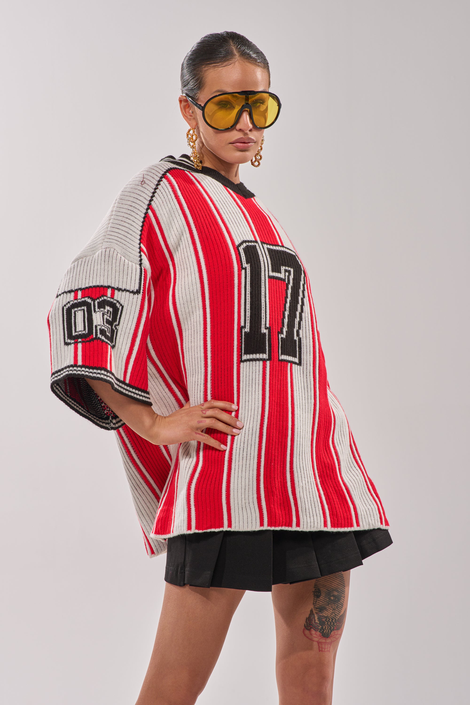 A person wearing the THROW SUM MO KNIT JERSEY TOP with "17" on the front and "03" on the sleeve, styled with large yellow sunglasses, bold gold earrings, a short black skirt, and a visible leg tattoo. 5