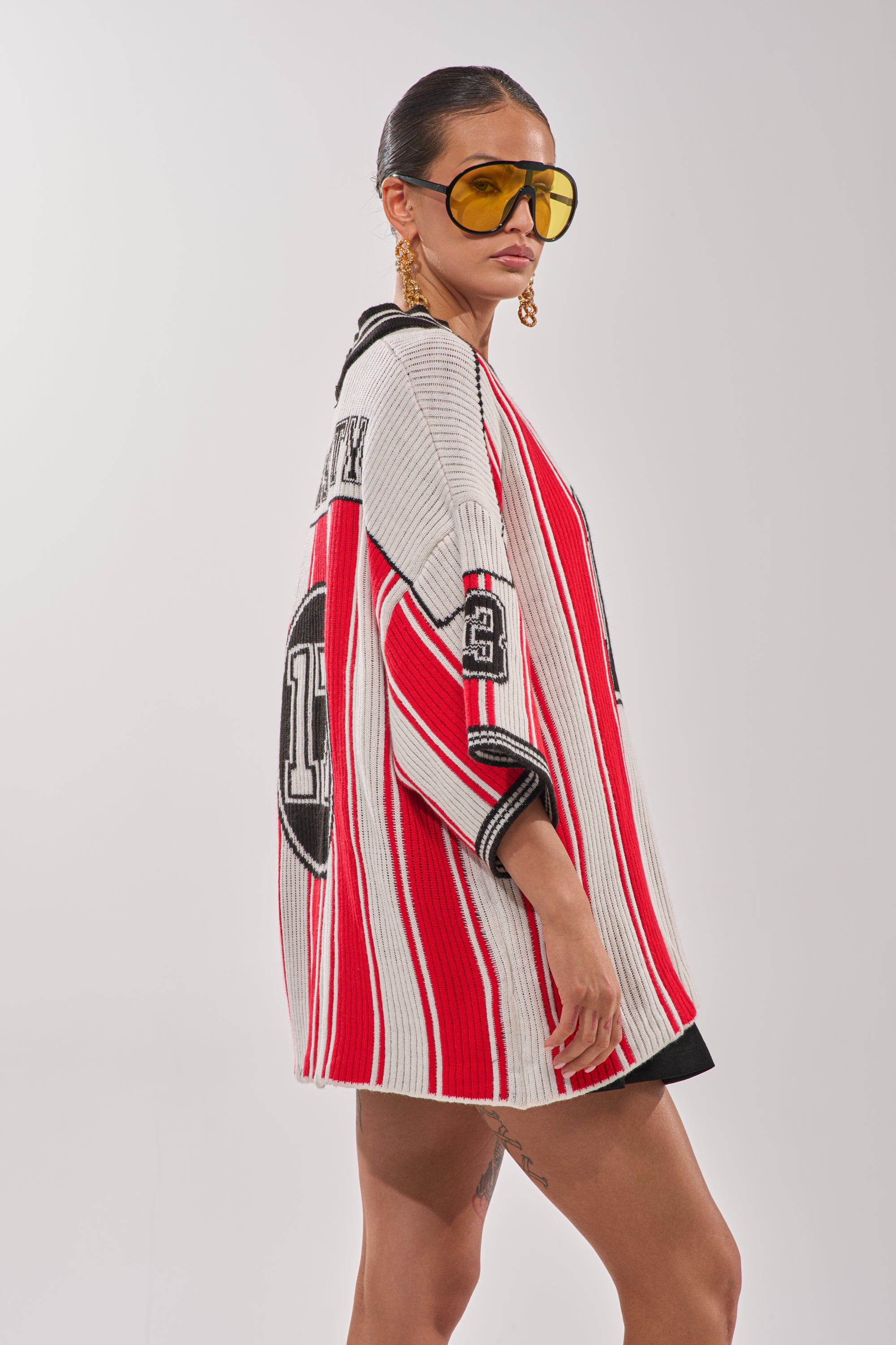 A woman in oversized yellow sunglasses and gold earrings poses in the THROW SUM MO KNIT JERSEY TOP, a varsity-inspired red, white, and black striped short-sleeve sweater with "13" on the back. Her hair is slicked back against a white background. 6