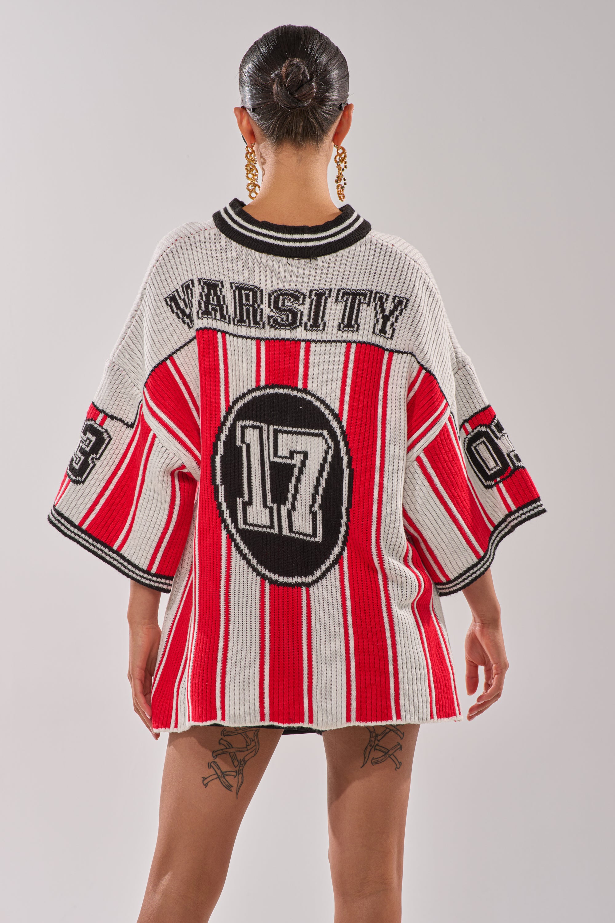 A person with slicked-back hair wears the THROW SUM MO KNIT JERSEY TOP, an oversized varsity-style sweater in red, white, and black featuring “VARSITY” and “17” on the back and “03” on the sleeves. They have visible leg tattoos and gold hoops. 7