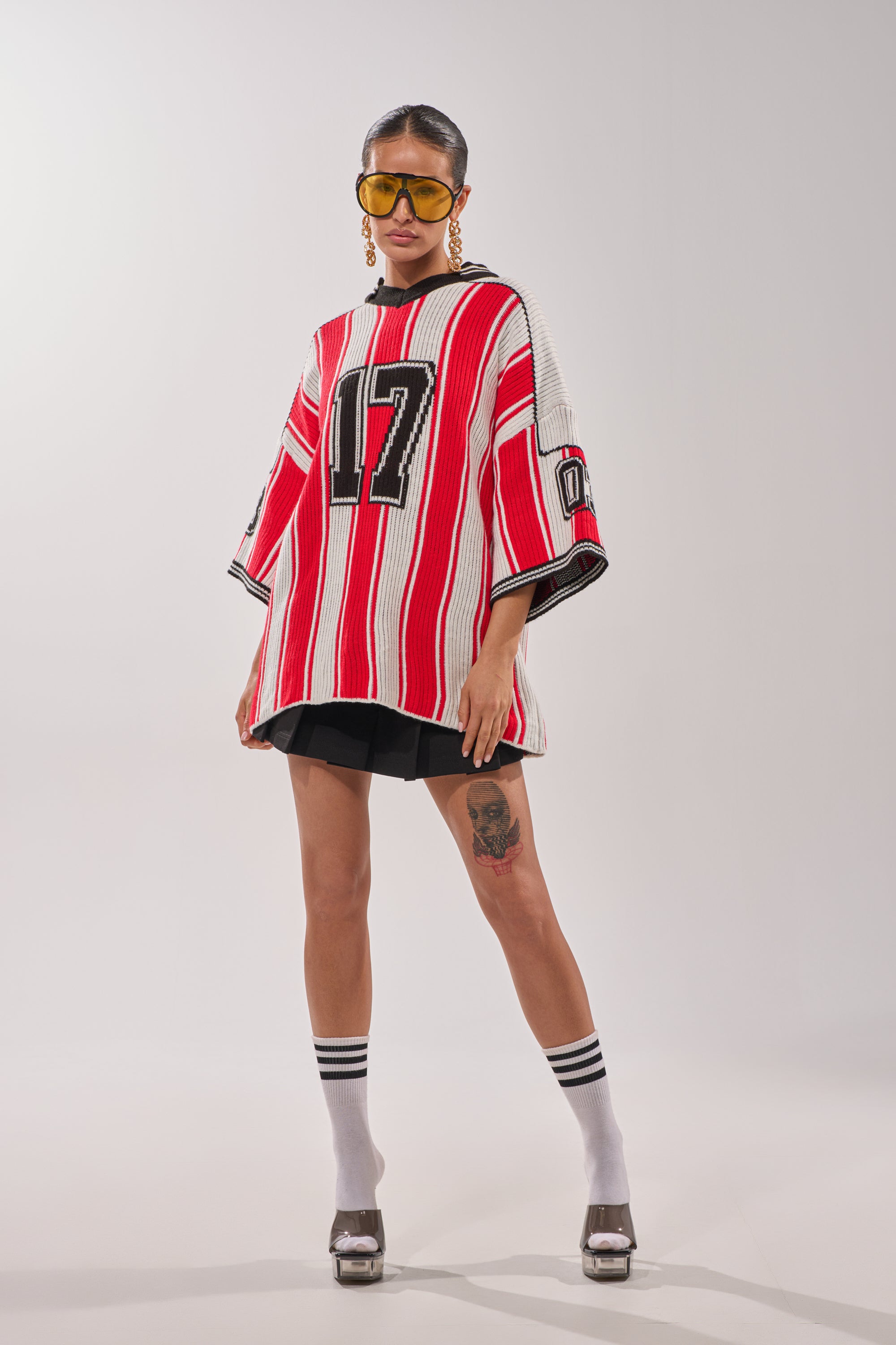 A woman stands confidently in the THROW SUM MO KNIT JERSEY TOP with red and white stripes and number 17, paired with black shorts, yellow sunglasses, platform heels, statement earrings, and a thigh tattoo. 1
