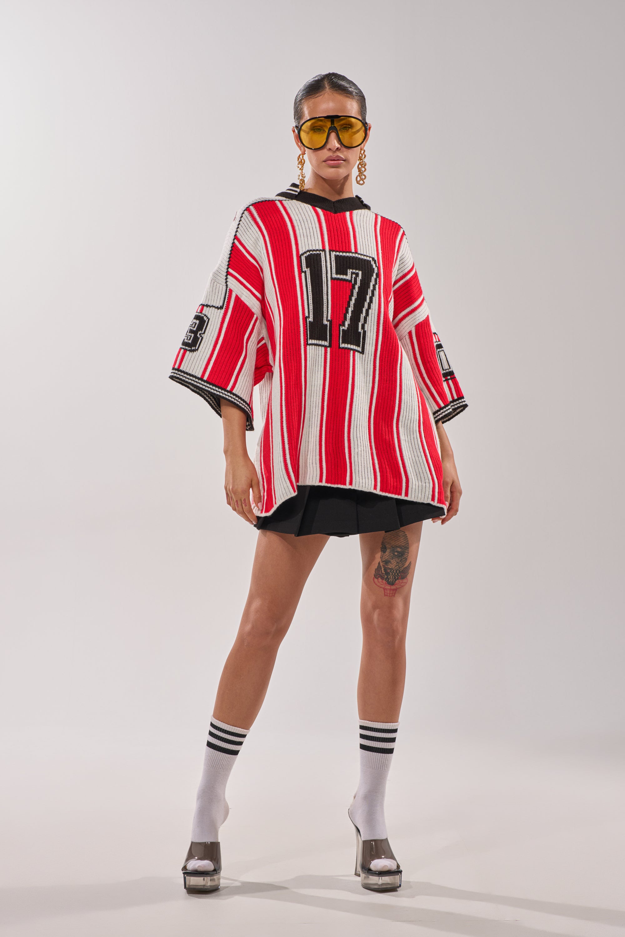 A woman poses in the THROW SUM MO KNIT JERSEY TOP, a red and white striped varsity sweater with "17," paired with black shorts, tall white socks with black stripes, silver platform heels, yellow sunglasses, and bold earrings. 0