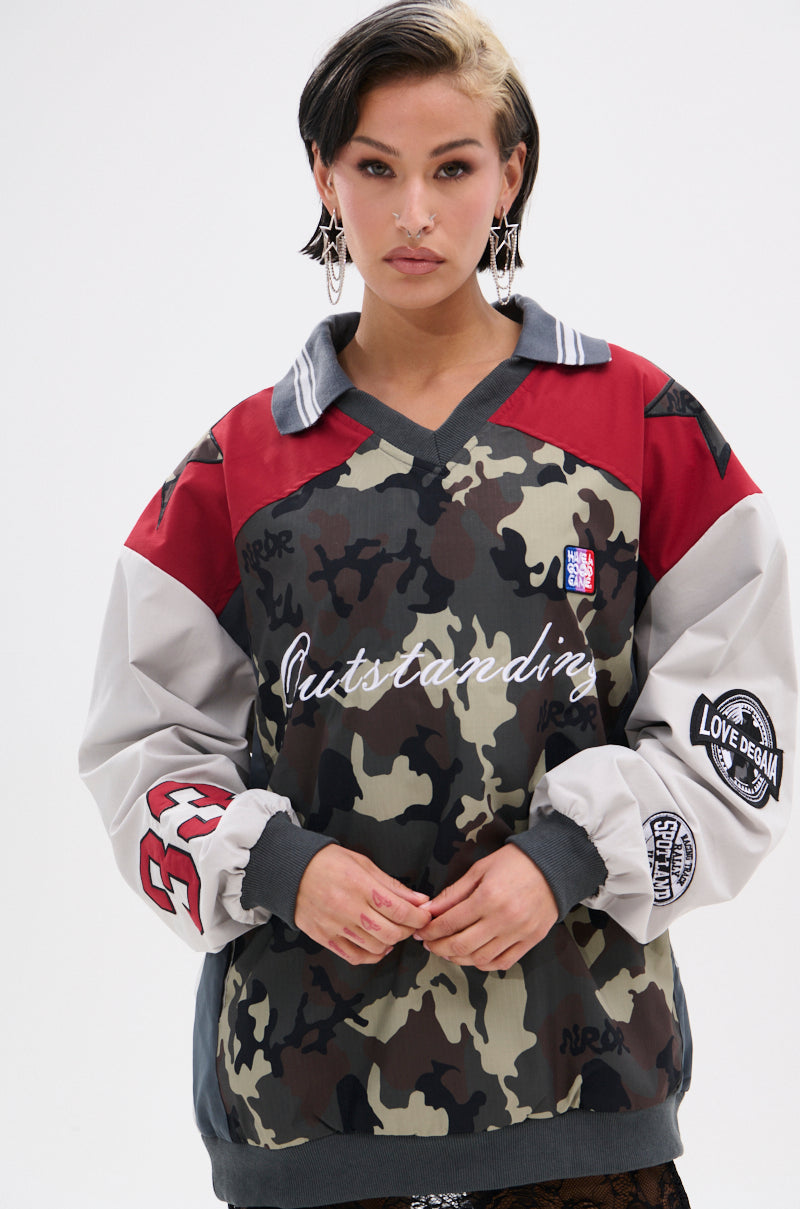 A woman with short dark hair wears the FLIP THE SWITCH PATCHWORK DETAILED JERSEY TOP, featuring a multicolored camo print with red and gray accents and “Outstanding” on the front, posing against a plain white background.