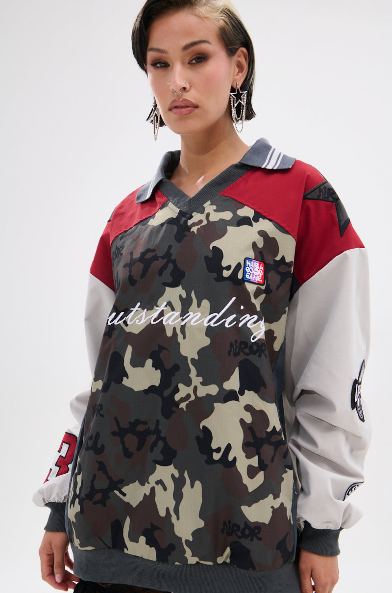 A woman with short dark hair wears the FLIP THE SWITCH PATCHWORK DETAILED JERSEY TOP, featuring an oversized relaxed fit, camo print, red shoulders, grey sleeves, bold patches, "outstanding" on the front, and statement earrings.