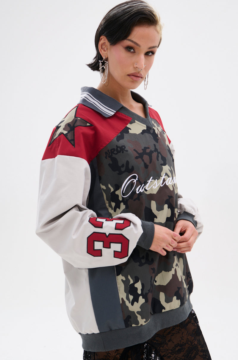A woman with short dark hair and hoop earrings wears the FLIP THE SWITCH PATCHWORK DETAILED JERSEY TOP, featuring a camo print, red star on the shoulder, and number 33 on the sleeve, standing against a white background.