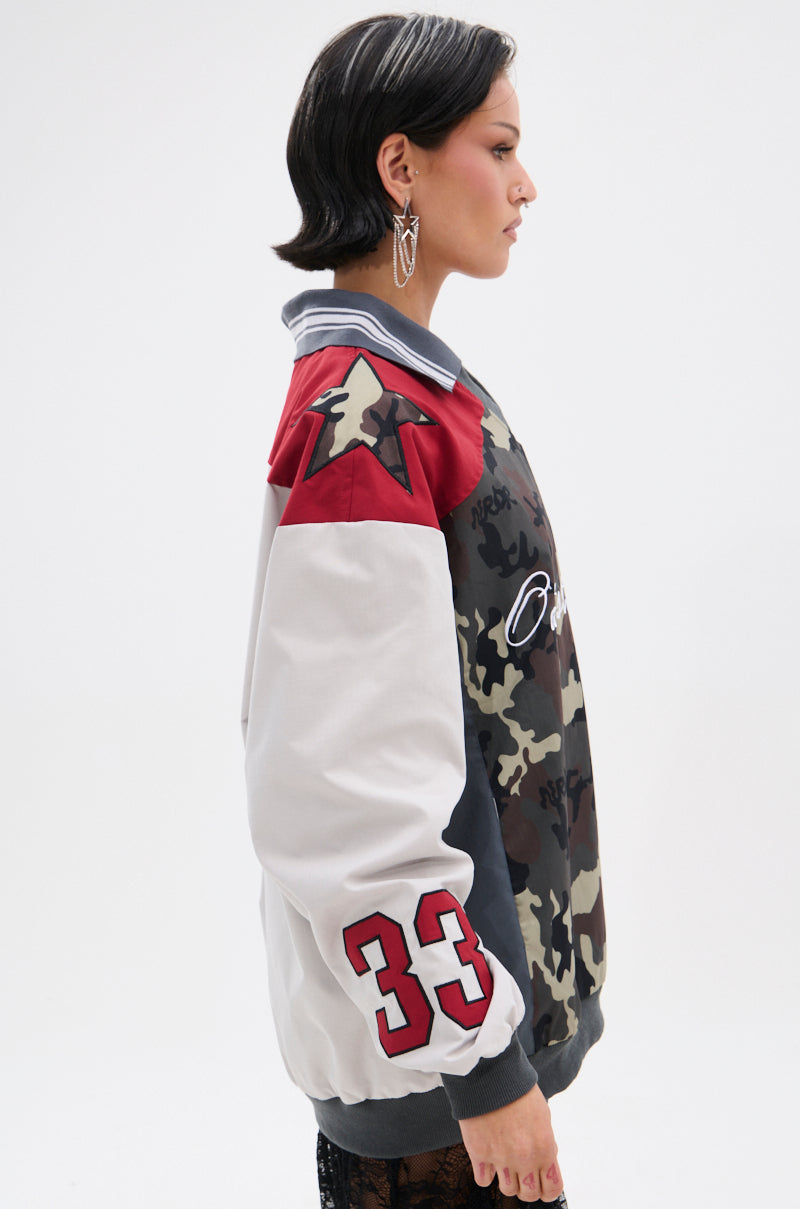 A person with short dark hair models the FLIP THE SWITCH PATCHWORK DETAILED JERSEY TOP, an oversized camo and white varsity jacket with a red star on the shoulder and number 33 on the sleeve, shown in profile against a plain white background.