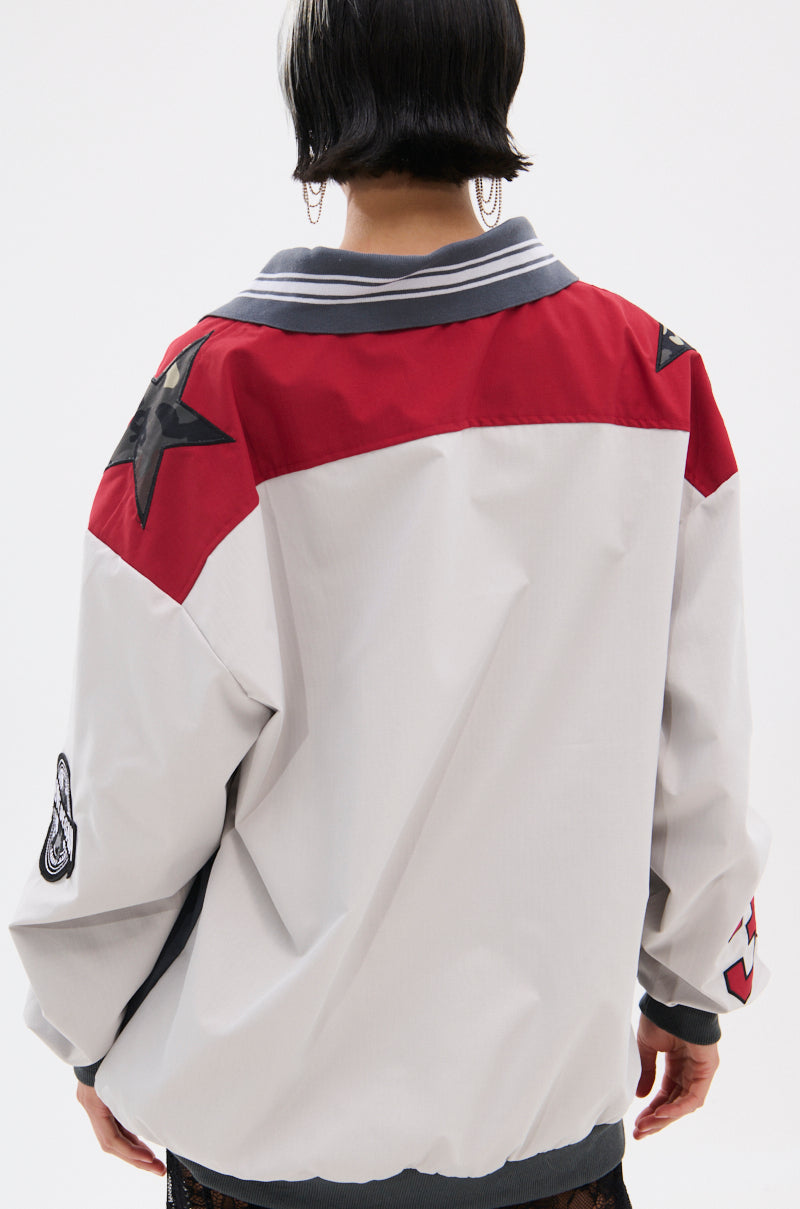 Person with short dark hair wears the FLIP THE SWITCH PATCHWORK DETAILED JERSEY TOP, an oversized light gray jacket with red accents, black star shoulder patches, and sleeve patchwork, shown from the back against a plain background.