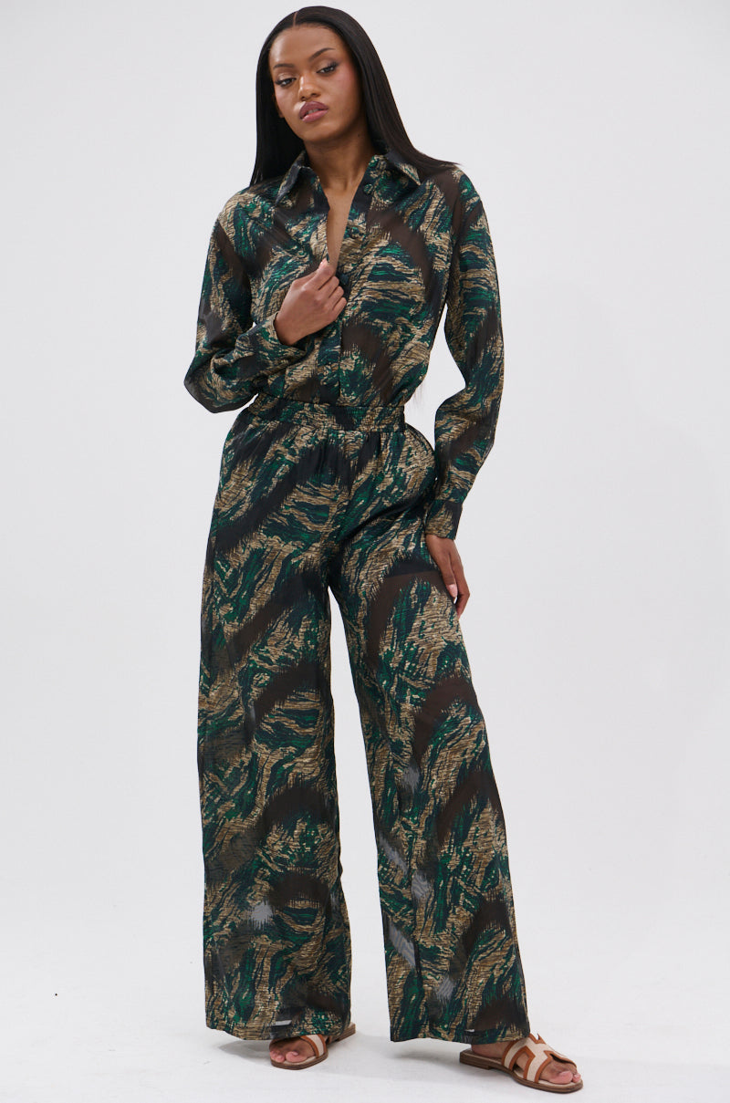 A woman stands confidently against a white background, wearing the IN TOO DEEP PALAZZO PANT featuring a green and brown abstract leaf pattern with a wide-leg silhouette, paired with straight hair and brown flat sandals.