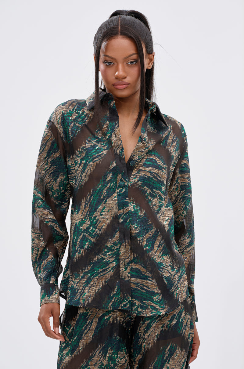 A woman wears the IN TOO DEEP BUTTON DOWN TOP, a sheer blouse with a green, black, and tan abstract pattern, paired with matching pants for an effortlessly stylish look against a plain light background.