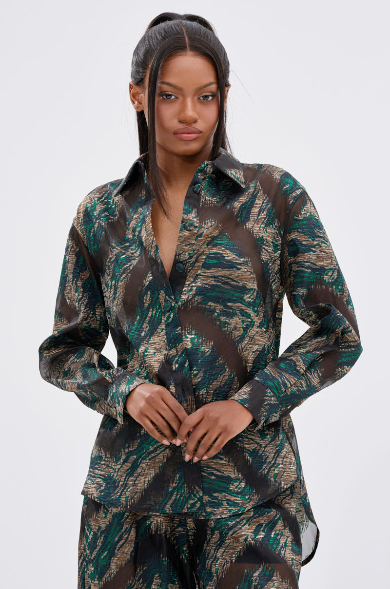 A woman with long, straight dark hair styled half-up confidently models the IN TOO DEEP BUTTON DOWN TOP, a sheer blouse featuring a green, black, and gold abstract pattern, against a simple light background.