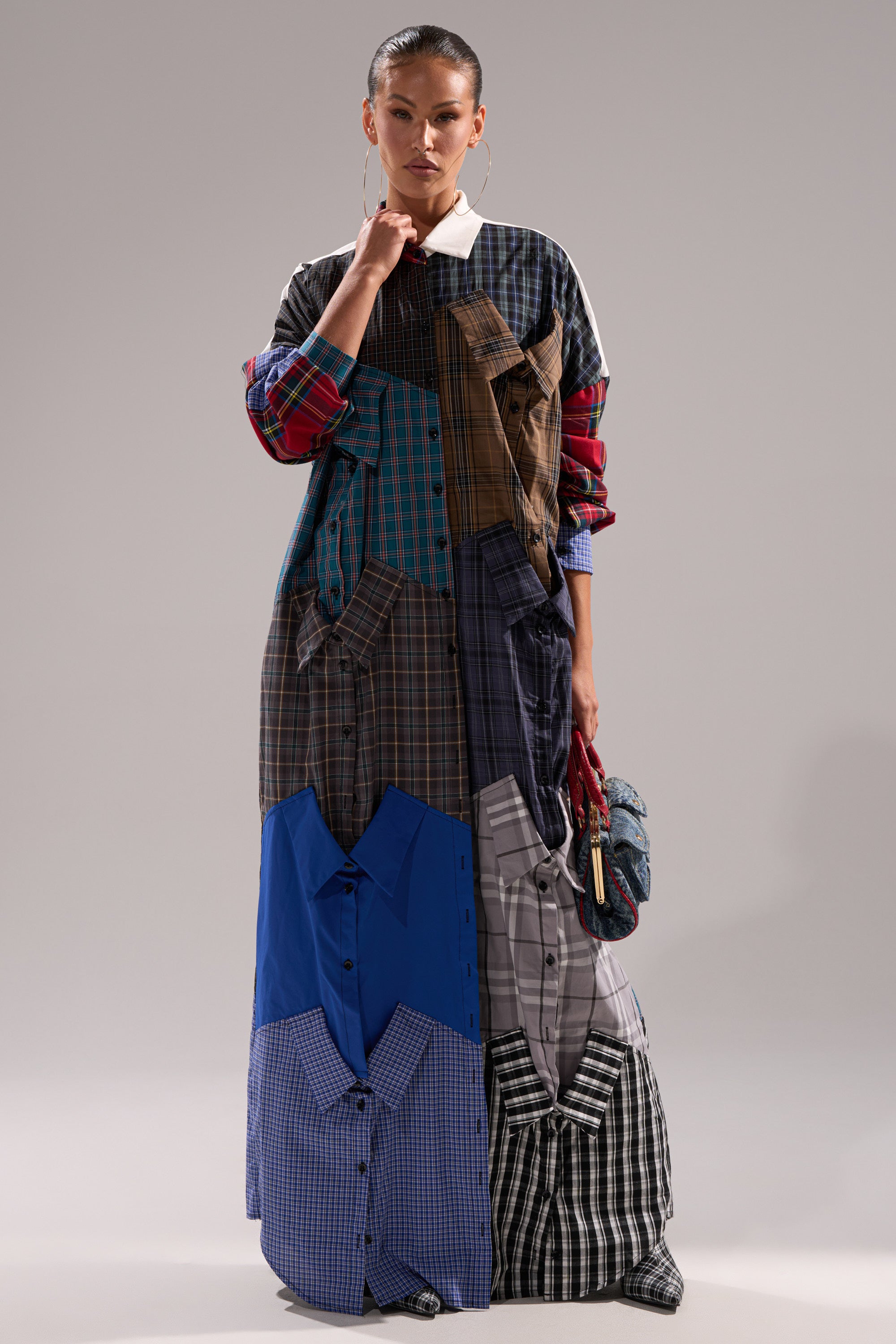 A woman wears the NOT YOUR BOYFRIENDS SHIRT DRESS, a vibrant plaid piece made from colorful, mismatched shirts, paired with a small blue purse as she poses against a plain background.