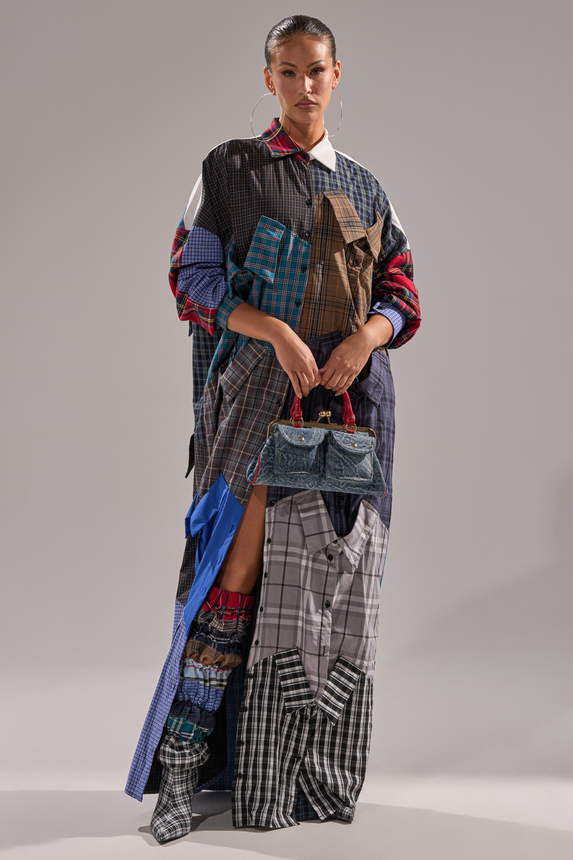 A model poses in the NOT YOUR BOYFRIENDS SHIRT DRESS, a long plaid shirt dress made from various checkered shirts, accessorized with a small denim handbag, patchwork boots, and large hoop earrings against a plain background.