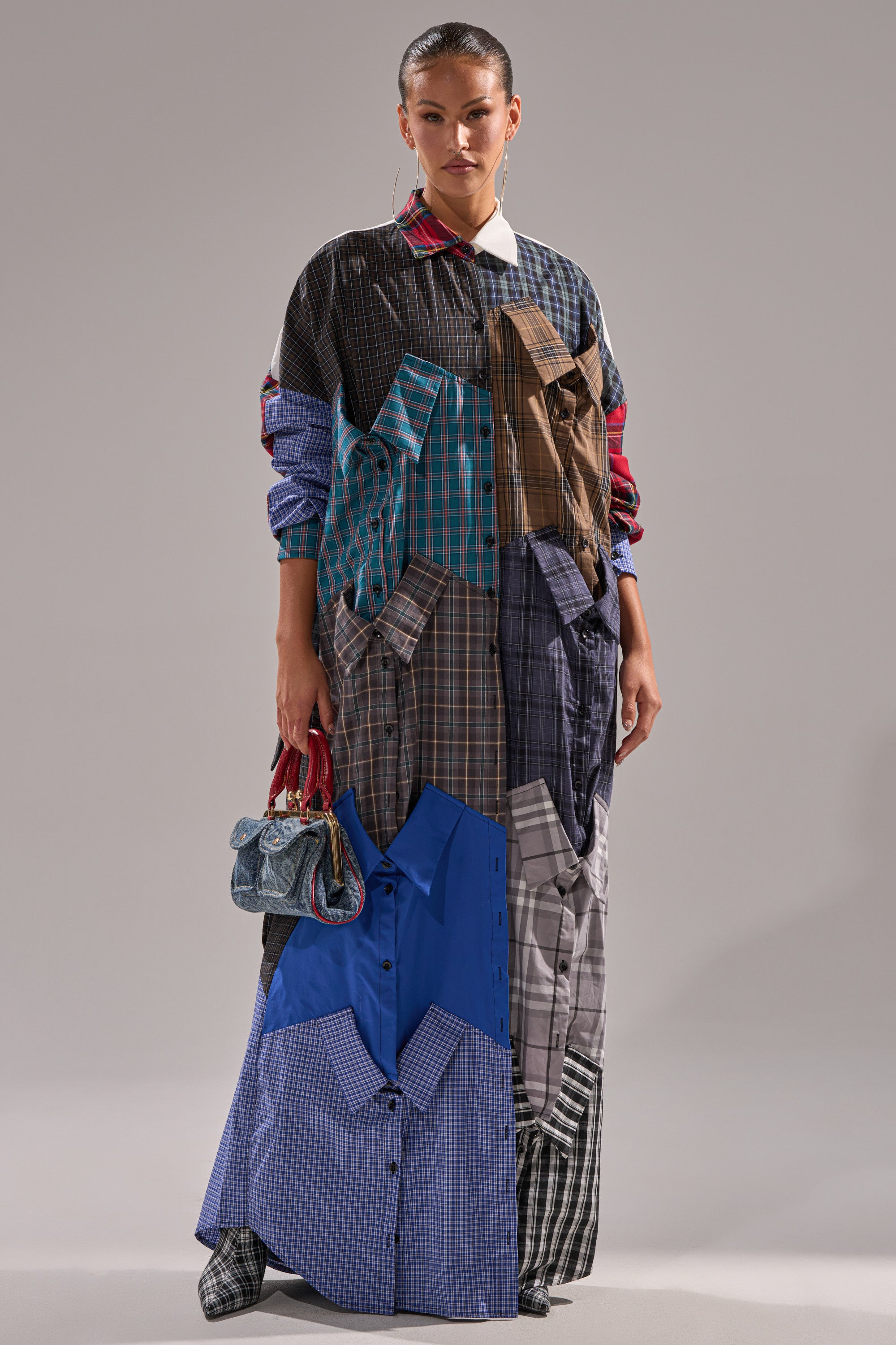 A woman poses against a gray background in the NOT YOUR BOYFRIENDS SHIRT DRESS, a maxi silhouette crafted from mismatched plaid and checkered fabrics. She holds a small textured handbag, with her hair pulled back.