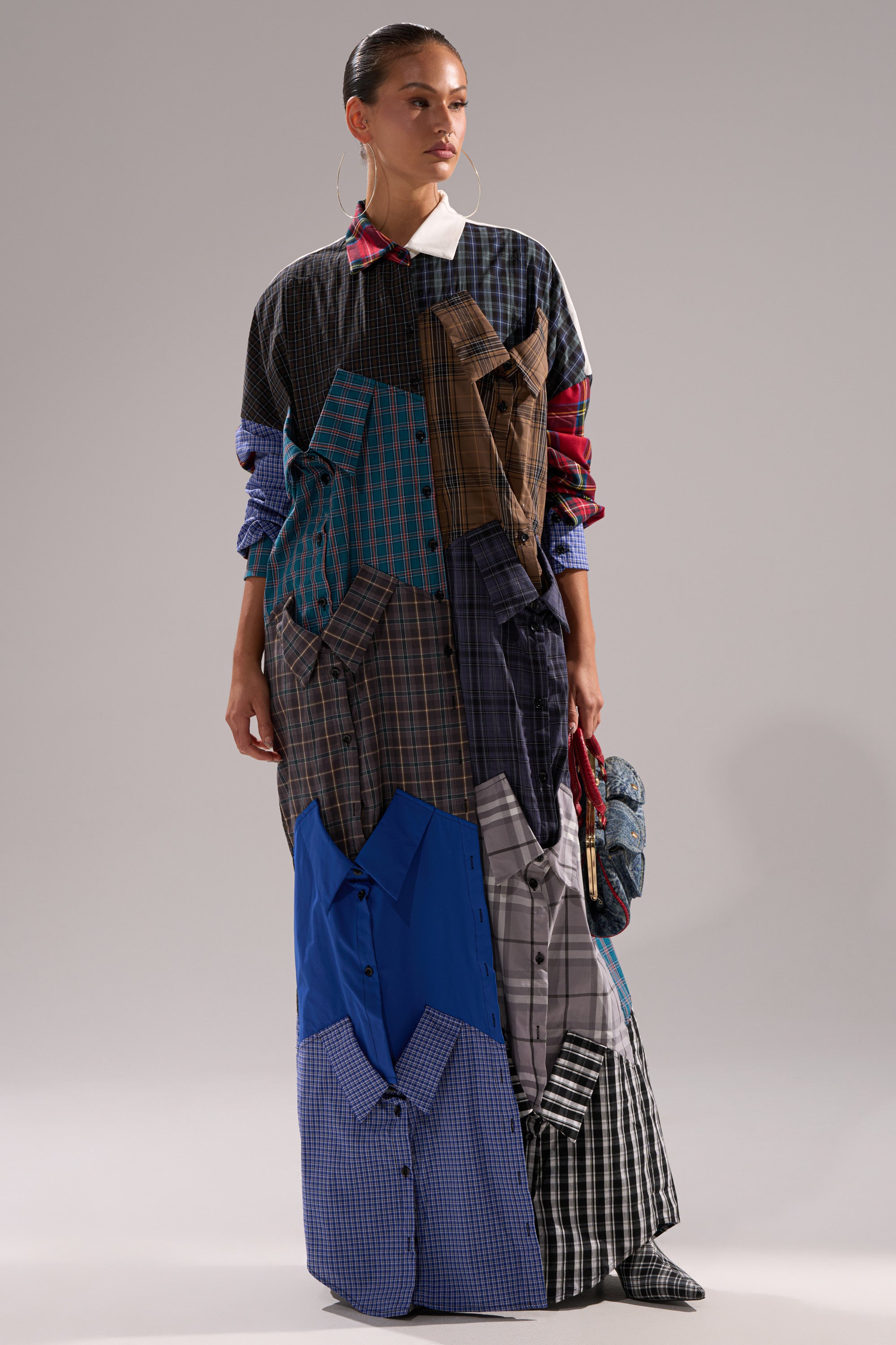 A woman wears the NOT YOUR BOYFRIENDS SHIRT DRESS—a long patchwork plaid dress made from repurposed shirts with rolled sleeves—paired with large hoop earrings and a small textured handbag.