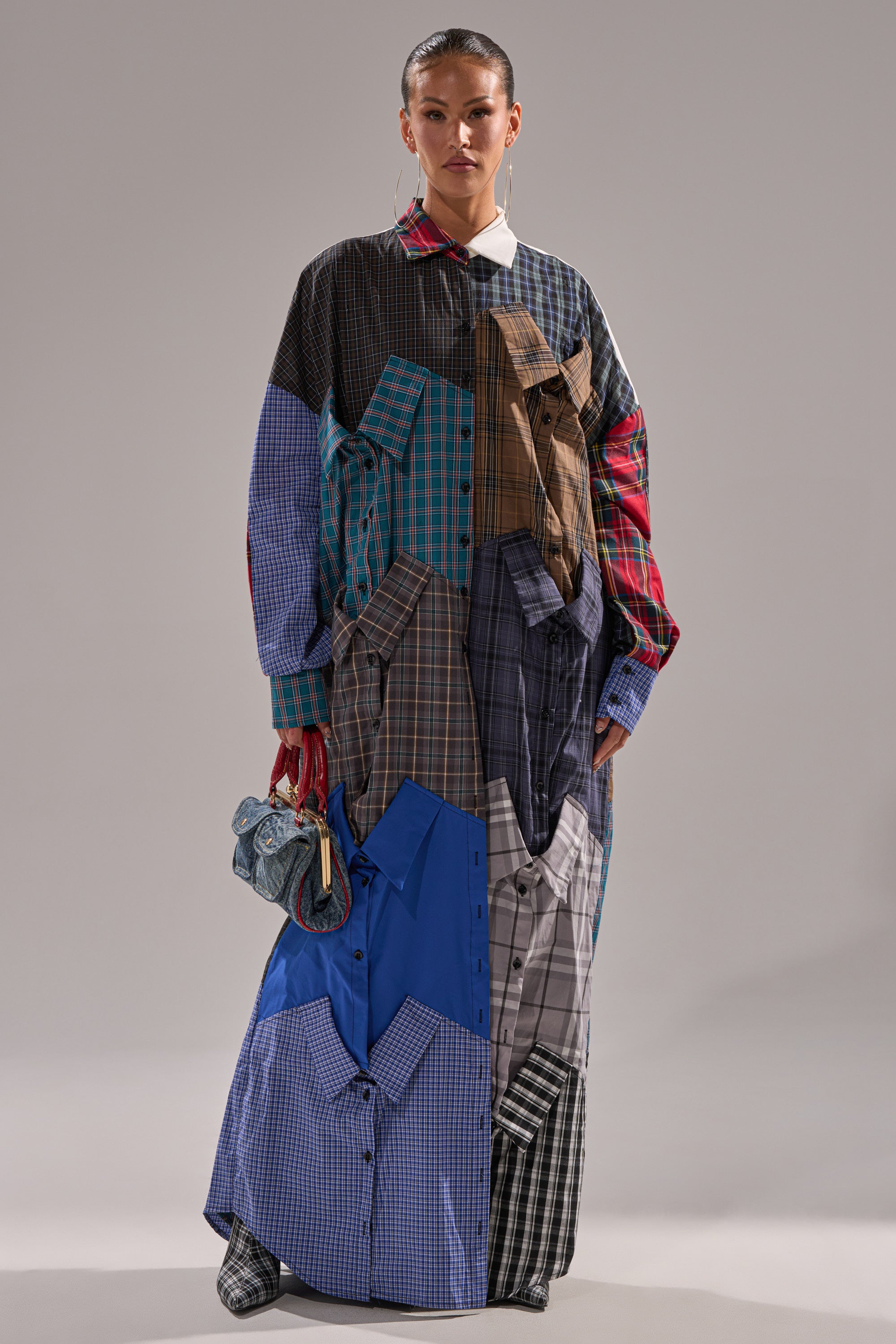 A woman models the NOT YOUR BOYFRIENDS SHIRT DRESS, a long, oversized dress made of mismatched plaid shirt patterns. She carries a small blue patterned handbag and wears her hair slicked back against a plain gray background.